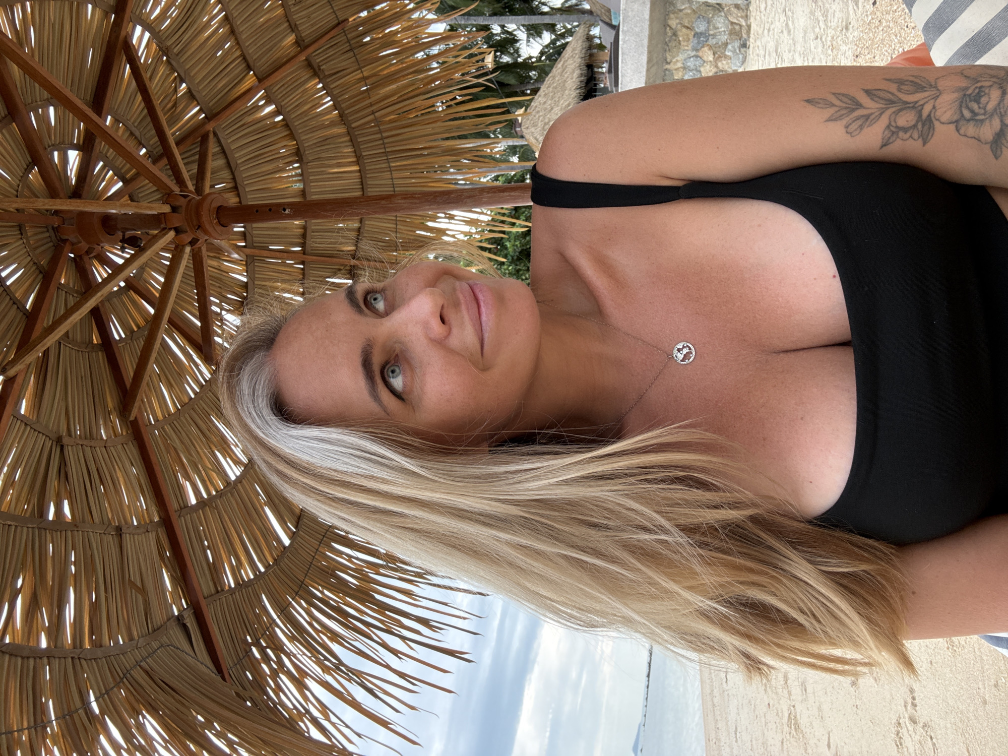 A medium shot shows a blonde woman with striking blue eyes sitting on a sandy beach under a thatched umbrella in the Ko Samui District, Thailand. She is wearing a black swimsuit and a silver pendant necklace. Her right arm, adorned with a floral tattoo, rests on her lap. The background reveals the ocean, a cloudy sky, and glimpses of resort buildings with thatched roofs. The sunlight filters through the umbrella, casting dappled shadows.