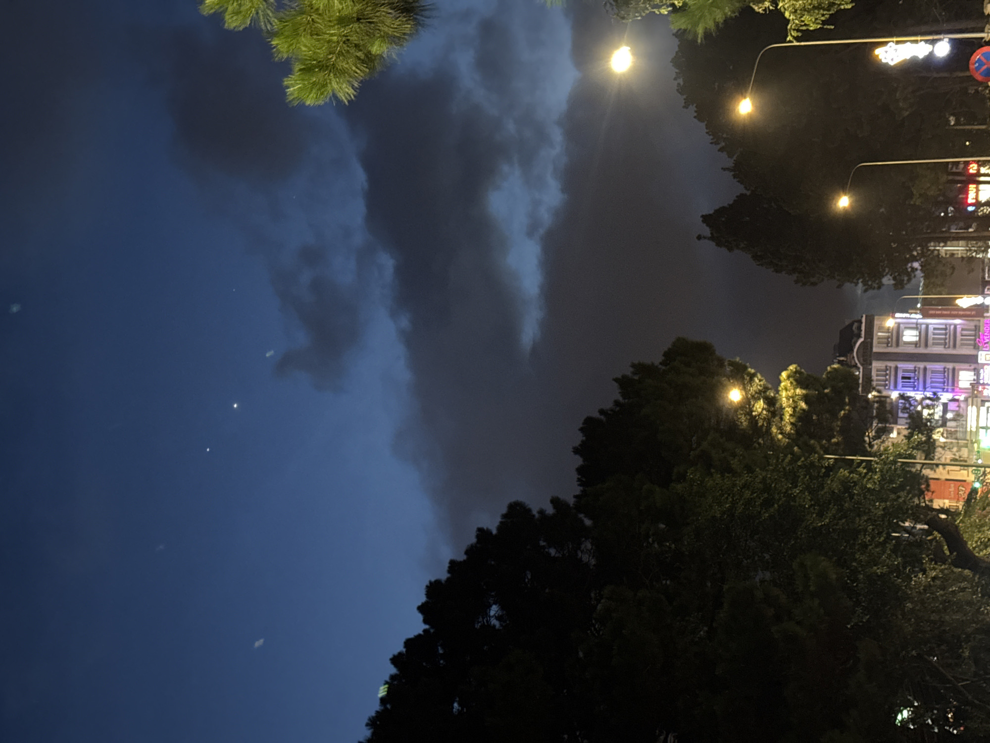 The image depicts a night scene in Da Lat, Vietnam, likely on a street lined with trees and buildings. The sky is a deep blue, with some dark clouds obscuring parts of it. A few faint stars or planets are visible. The foreground is dominated by the dark silhouettes of trees, with some illuminated by streetlights. On the right side of the image, a bright streetlight casts a warm glow.
In the mid-ground, several buildings are visible, their windows illuminated by interior lights, giving them a warm and inviting appearance. Some of these buildings appear to be residential or commercial establishments. Neon signs and other forms of artificial lighting are present, contributing to the urban ambiance. The street itself is not clearly visible, but the presence of streetlights and buildings suggests a well-trafficked area.
The overall impression is one of a quiet urban night, with the natural elements of trees and sky contrasting with the man-made structures and lighting. The image captures the unique charm of Da Lat at night, a city known for its cool climate, pine forests, and French colonial architecture.