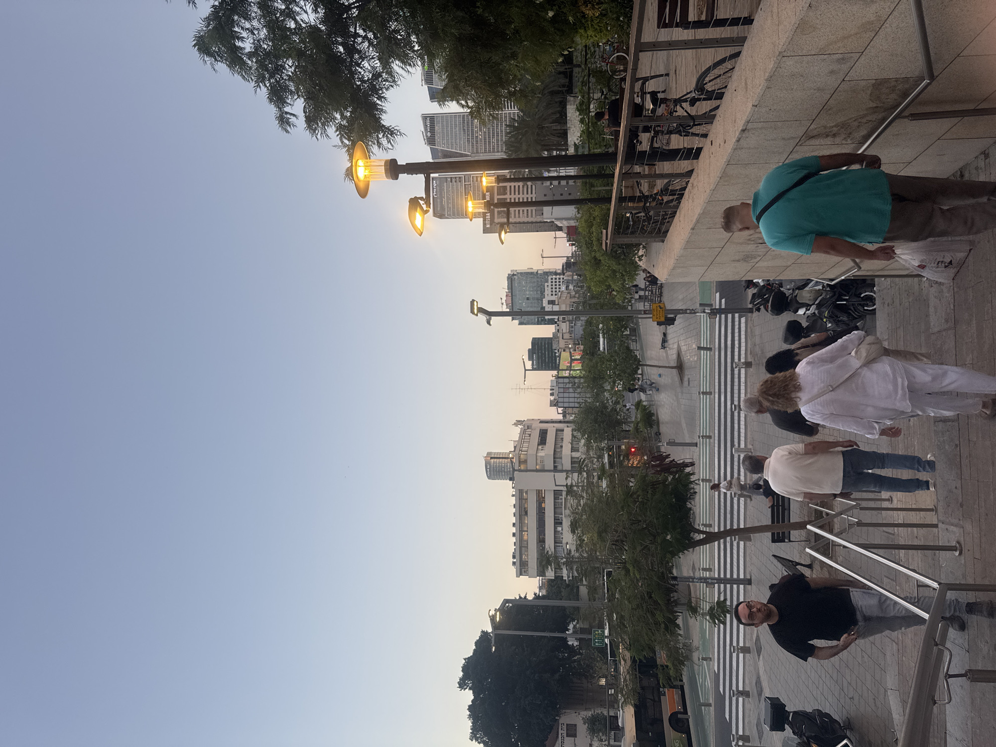 The image captures an urban street scene in Tel-Aviv, Israel, during what appears to be early evening or twilight. The sky transitions from a light blue at the top left to a pale orange-yellow horizon line, indicating the time of day, and streetlights are brightly illuminated.
In the foreground, several people are visible on a paved sidewalk and a set of stairs or a ramp with metal railings. On the far right, a man in a teal/turquoise shirt and khaki pants walks up the stairs, holding a white bag. Below him, a woman with light hair in a white top and light pants is seen, along with another man in a light-colored polo shirt and blue jeans. At the bottom of the stairs, a man in a black t-shirt and grey jeans, wearing glasses, is looking towards the viewer while holding a phone.
The street is bustling with activity and infrastructure. Multiple streetlights with distinctive yellow-orange glow line the sidewalks. Parked bicycles are visible in racks, and motorcycles/scooters are parked on the pavement. Trees and lush green foliage, including some palm trees and larger trees, are interspersed throughout the scene, offering a contrast to the urban architecture.
In the mid-ground and background, modern high-rise buildings and other commercial structures form the city skyline. On one prominent building, the text "NISIM BER" and partially visible "ARCHI" (likely for architects) can be read. A pedestrian crosswalk (zebra crossing) is clearly marked on the street, and traffic lights are also present. A bus or large vehicle is partly visible in the lower left corner, bearing the text "ING". The overall atmosphere suggests a lively city environment transitioning from day to night.