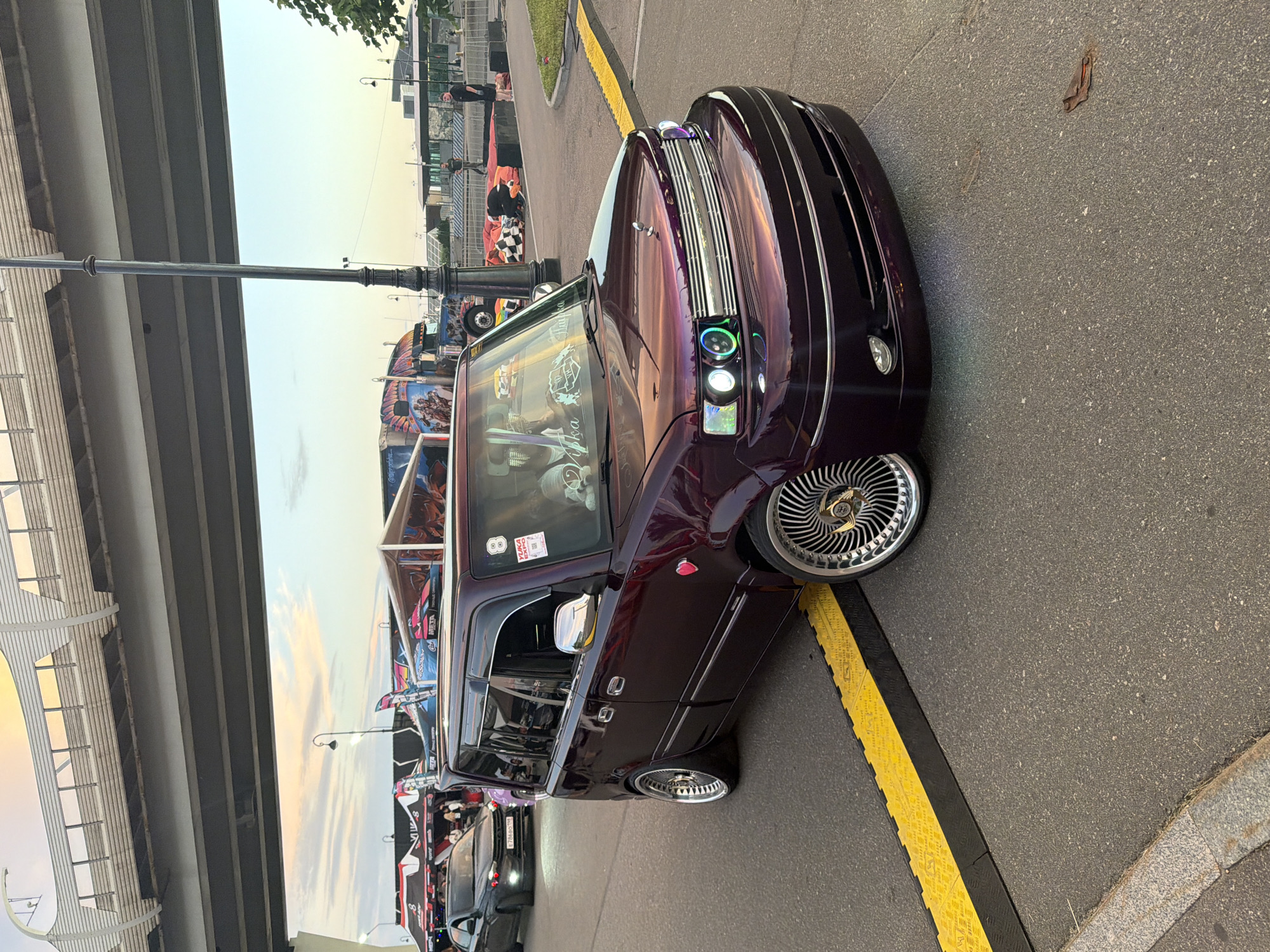 A modified, dark purple Toyota bB is parked on an asphalt surface, facing right. The car has a custom front bumper, chrome grille, and aftermarket headlights with multi-colored halos. It is equipped with large, chrome multi-spoke wheels. A yellow and black speed bump is positioned in front of the car. In the background, several tents with colorful graphics are set up, and a dark-colored sedan is visible further down the road. A bridge structure spans the upper portion of the image. The sky above is a mix of soft yellow, orange, and blue, indicating either sunrise or sunset.