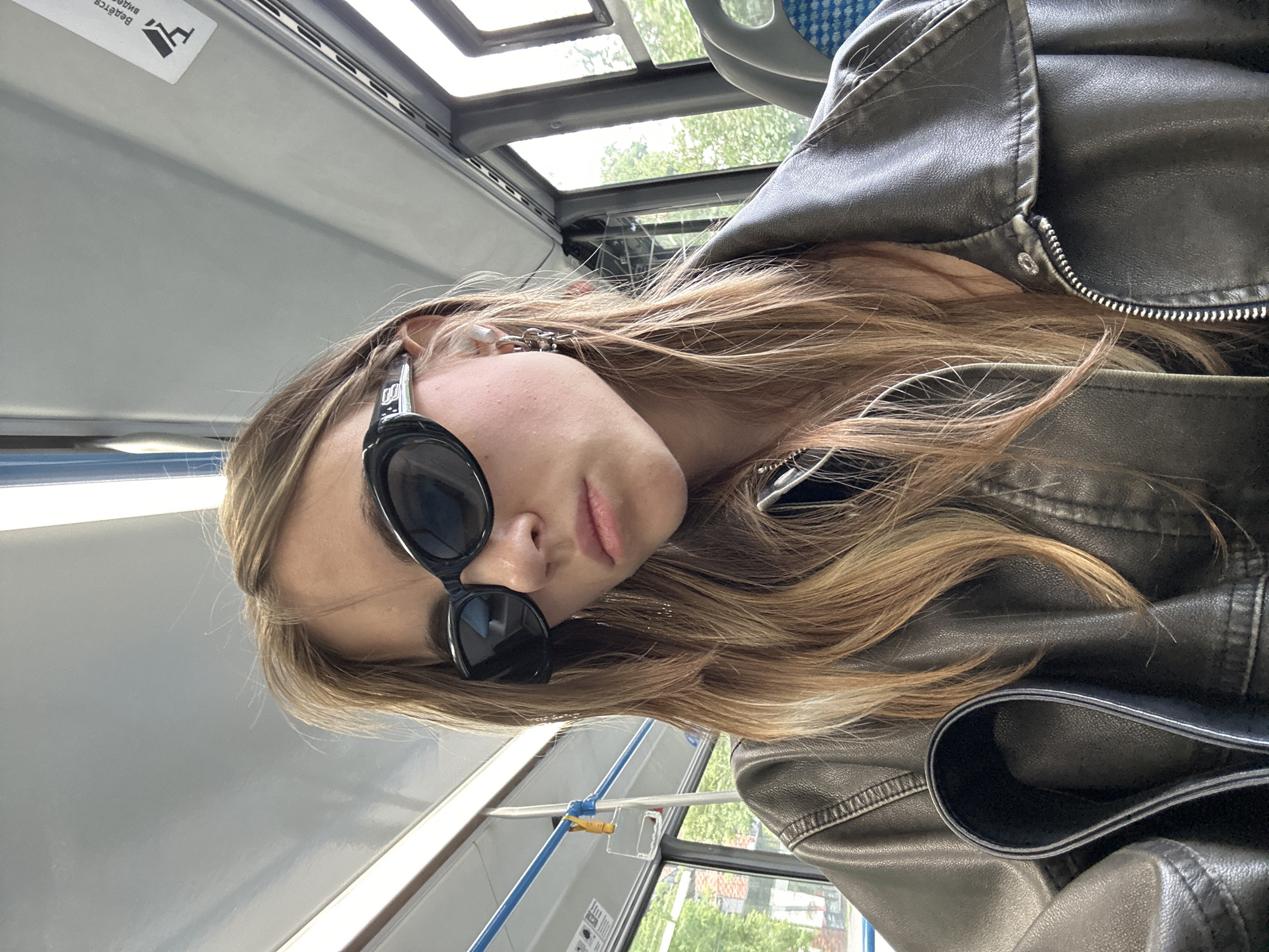 A young woman wearing sunglasses and a leather jacket is sitting on a bus. She has long, light brown hair and is wearing large, black, oval-shaped sunglasses. She also has a silver earring in her visible ear, which appears to be a chain-link design. The interior of the bus is visible behind her, with grey ceilings, blue handrails, and windows showing greenery outside. The overall impression is a candid shot taken from a close-up perspective, focusing on the woman and her fashion choices within the public transportation setting. The image does not provide explicit details that would allow for a definitive location in Moscow, Russia.