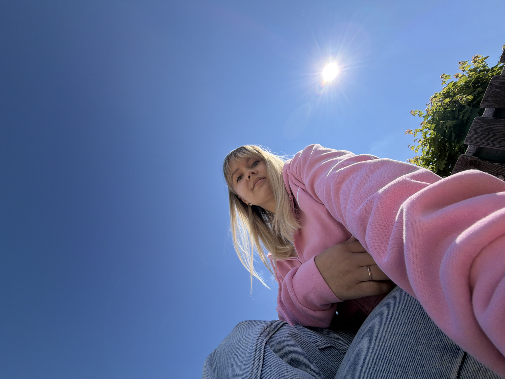 This outdoor photograph, taken in Chaikovsky, Russia, depicts a young woman from a low-angle perspective, looking down towards the camera. She has blonde hair with bangs and is wearing a light pink hooded sweatshirt and blue denim jeans. A plain golden ring is visible on the fourth finger of her left hand, which is resting near her hip.
The setting is bright and sunny under a clear, expansive blue sky. The sun, prominent in the upper right portion of the frame, creates a strong lens flare. To the right, a lush green tree or bush and a wooden structure, possibly a fence or part of a bench, are partially visible. The majority of the image is dominated by the clear blue sky, emphasizing the low camera angle. There is no visible text in the image. The time of day appears to be daytime, given the intense sunlight.