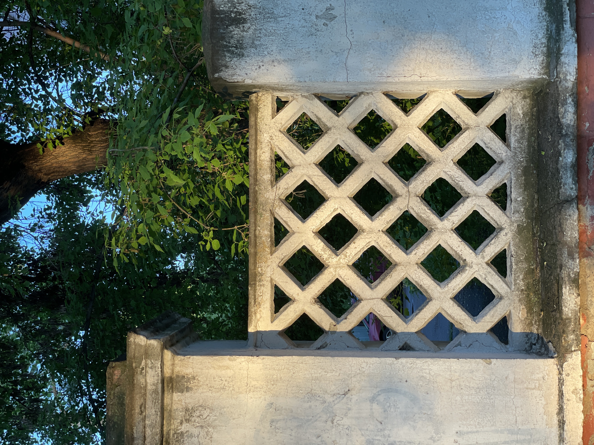 A low-angle, rotated view shows a weathered, rectangular, white concrete lattice wall structure with diamond-shaped openings. Behind the wall, green trees and blue sky are visible. To the right, a red drain pipe runs vertically along the wall. The structure is presumably located in Zheleznogorsk, Russia.