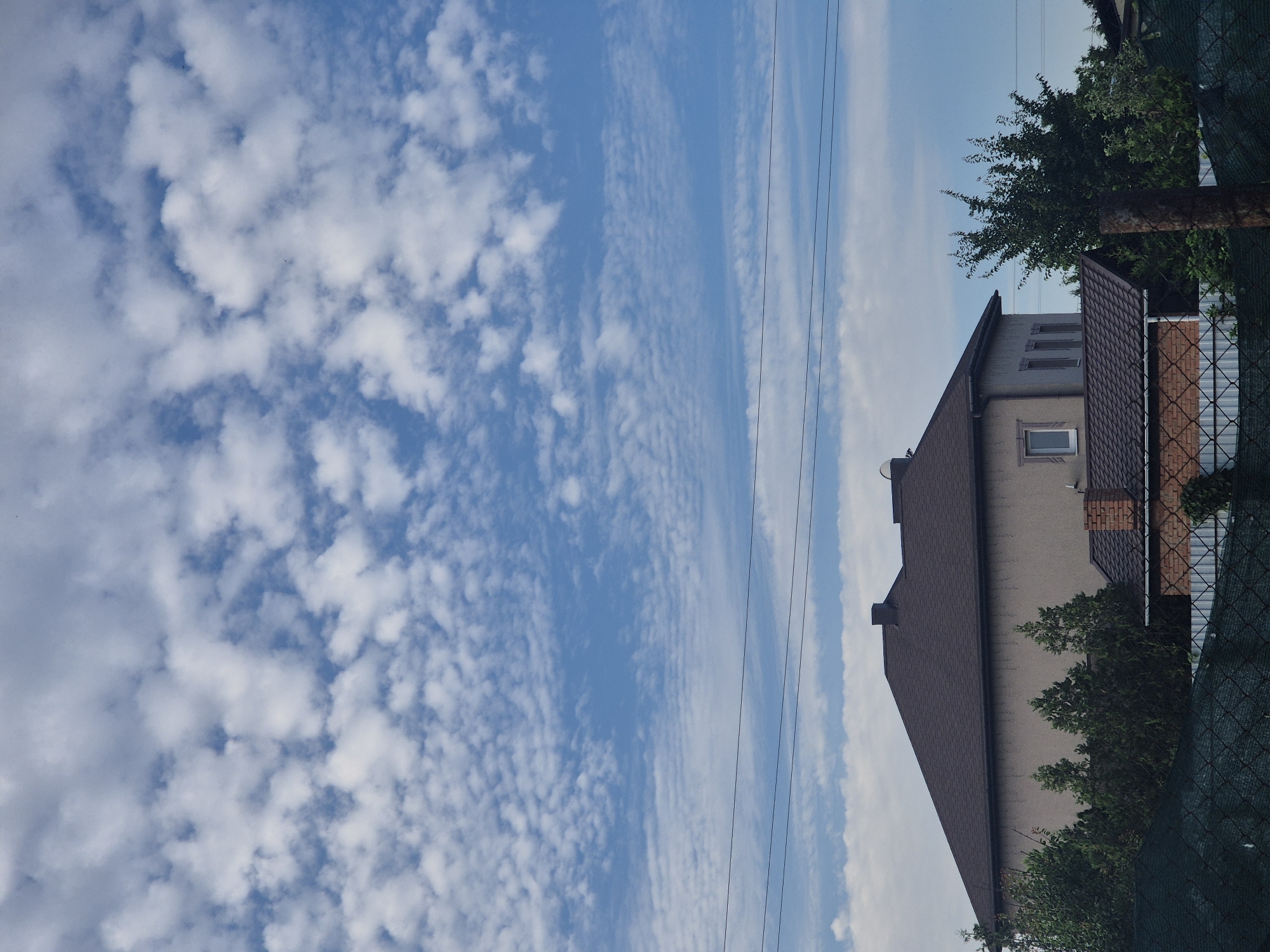 In Sozonivka, Ukraine, a view captures a residential area under a dynamic sky. The upper portion of the image is dominated by a bright blue sky filled with scattered, puffy white clouds. Below the sky, three parallel dark electrical wires traverse horizontally across the frame. In the middle ground, the roofline of a multi-story building with a dark brown, shingled roof is visible. The building's facade is a neutral, light beige color, with a single rectangular window prominently placed on the side facing the viewer. To the right of this building, a smaller structure with a darker brown roof and a brick chimney is partially visible. A dense line of green trees and foliage partially obscures the lower portions of the buildings. In the foreground, a dark green mesh fence runs horizontally across the bottom of the image, with a rusty metal pole standing vertically on the right side, partially in front of the fence. The scene depicts a quiet, domestic setting with elements of infrastructure and natural surroundings.