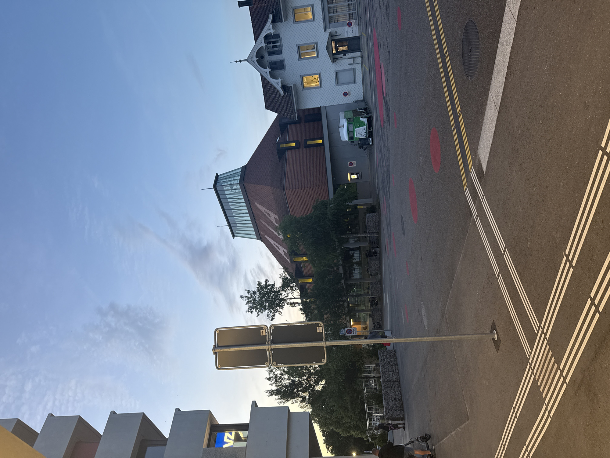 This media file captures an evening street scene in Liestal, Switzerland, rotated 90 degrees clockwise from its natural orientation. The sky, seen on the left (top when correctly oriented), is a clear, light blue with faint wispy clouds, indicating either dusk or early evening, a time reinforced by the illuminated windows in several buildings.
The setting is an urban street flanked by diverse architecture. On the far right (top when correctly oriented), stands a charming, traditional white building with a patterned dark roof and decorative gables, its windows glowing with warm light. Adjacent to it, closer to the center, is a larger building featuring a reddish-brown tiled roof and a distinctive modern glass and metal section, likely a skylight or roof extension. This building also has illuminated windows. Further to the left (bottom when correctly oriented), modern architectural elements with sharp, angular designs are visible. One such structure displays glowing blue letters, possibly "VZ" or "VZA," indicating text on a sign or display.
The paved asphalt road occupies a significant portion of the foreground and center. It features clear road markings, including double solid yellow lines and distinct triple white lines, possibly indicating a pedestrian crossing or a specific lane. Scattered red circular markings are also present on the asphalt. A circular manhole cover is embedded in the road surface. A light-colored curb separates the road from a sidewalk or pedestrian area.
Several trees with green foliage are interspersed throughout the scene, adding natural elements to the urban landscape. A prominent metal pole with two dark rectangular signs stands in the center-left. Near the traditional white building, a small green and white utility vehicle is parked. In the lower-left corner, two figures are visible on the sidewalk, one wearing a dark shirt and shorts, standing near a black electric scooter. A low wall constructed from gabions (wire cages filled with stones) is also present, acting as a boundary or decorative feature.
The image presents a calm, everyday snapshot of a street in Liestal during the transition from day to night.