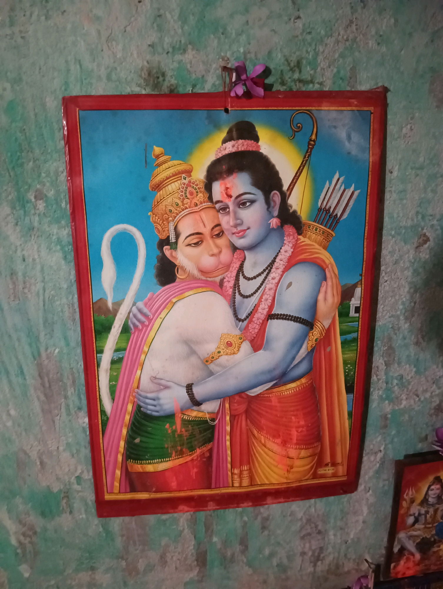 The image displays a framed painting of two figures, likely deities from Hindu mythology, on a textured, light green wall. The central figures are a blue-skinned individual wearing a crown and adorned with necklaces and bracelets, embracing a monkey-like figure with a crown and a pink garment draped over its shoulders. The monkey figure also has a long, white tail curling upwards. Behind the blue-skinned figure, a quiver of arrows is visible. The painting is bordered by a red frame. To the right of the main painting, a smaller framed image shows a seated figure with multiple arms, holding a trident, on a flaming base. The overall setting appears to be an indoor wall, possibly in a residential or religious space in Dakhin Parsa, India, given the context provided. The wall surface is rough and shows signs of wear.