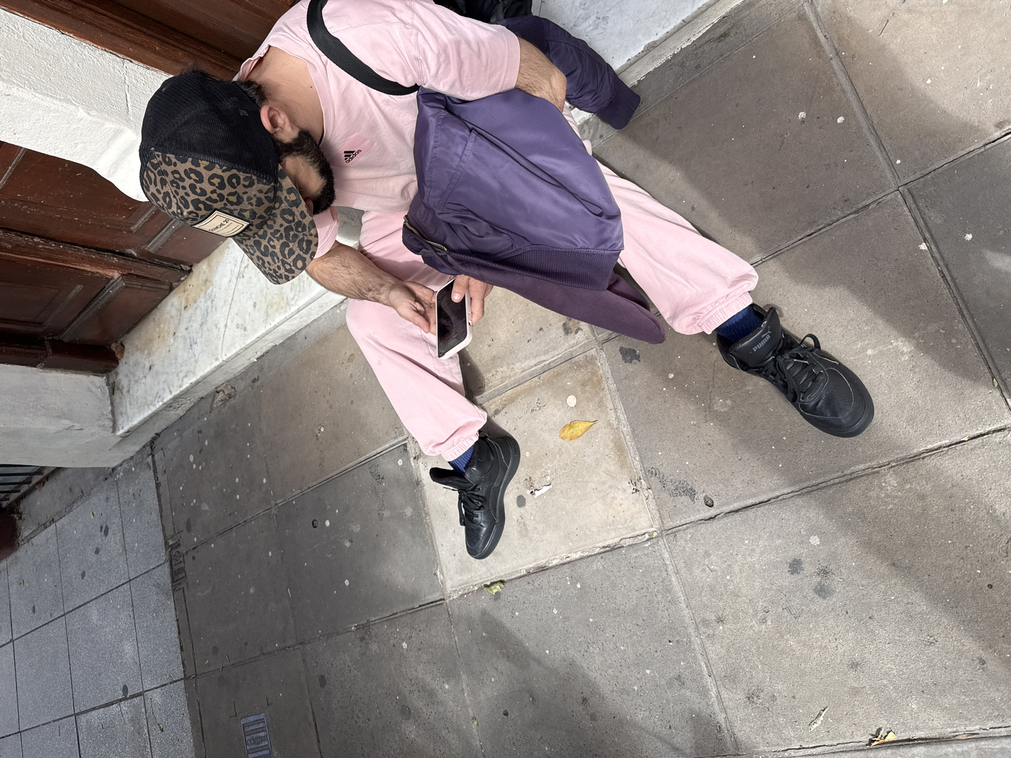 An overhead shot captures a person sitting on a concrete sidewalk next to a building, likely in Buenos Aires, Argentina. The individual, who has a dark beard, is dressed in a light pink long-sleeved shirt featuring an "adidas" logo and matching pink track pants. They are wearing a black mesh-back cap with a leopard print brim, which has a small brown label with "Cracker 21" printed on it. Black athletic shoes with a visible "Puma" logo and blue socks complete their attire. A dark purple jacket or bag is draped over their left arm and lap.
The person is holding a white smartphone in both hands, their gaze directed downwards at the screen, indicating they are engaged with the device. They appear to be leaning against a dark wooden door or lower part of a building entrance, which features a white stone ledge or trim above. The sidewalk is paved with square concrete slabs, showing some dirt, cracks, and scattered light specks. Strong, distinct shadows are cast across the ground, suggesting it is a sunny daytime. A single yellow leaf lies on the sidewalk near one of the person's feet.