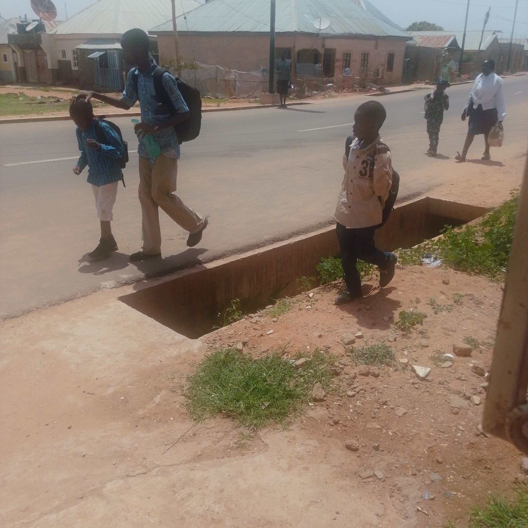 The image captures a street scene in Gombe, Nigeria, under what appears to be a bright, sunny day, suggesting it is mid-day. The foreground is dominated by a dry, dusty earth path with scattered rocks and patches of dry grass. A drainage ditch runs along the edge of the paved road.
On the paved road, three children are visible, along with one adult. In the foreground, a young boy in a light-colored, collared shirt with the number "31" and text partially visible beneath it, walks away from the camera towards the right. He wears dark pants and dark shoes and carries a dark backpack.
To the left of the frame, two other individuals, likely older boys or young men, are walking on the road. The one in front, closer to the camera, wears a blue checkered shirt, white pants, and dark shoes, carrying a dark backpack. The person behind him, wearing a blue checkered shirt and khaki pants, has his hand on the head of the boy in front. He also carries a dark backpack and holds a green object and a light-colored bottle.
Further down the road, a person in a military-style camouflage uniform with a backpack is walking towards the foreground. To their right, a woman in a white top and dark skirt walks away from the camera, carrying a light-colored bag over her shoulder.
The background shows a street lined with buildings, some appearing to be houses with corrugated metal roofs. The overall scene depicts people, likely students or pedestrians, moving along a street in an urban or semi-urban environment in Nigeria. The atmosphere seems ordinary, with no overt signs of distress or celebration.