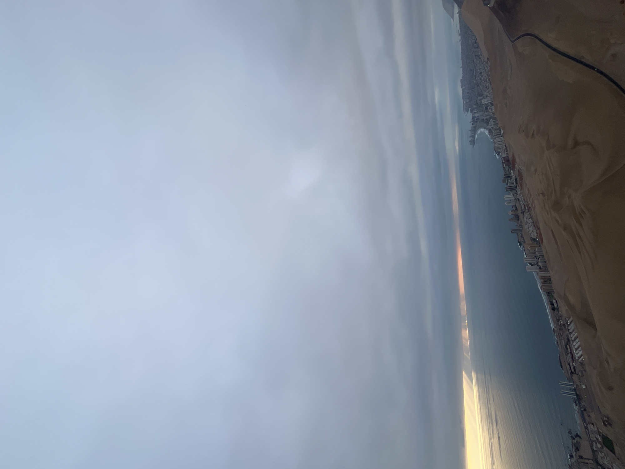 The image depicts a coastal scene, with a city lining the shoreline of an ocean or large body of water. Buildings, presumably high-rises, are visible along the beach, adjacent to what appears to be a sandy area, possibly dunes. A road runs parallel to the coastline. The sky is overcast, with a diffuse light possibly indicating sunrise or sunset. The water reflects this light. The location is unknown.