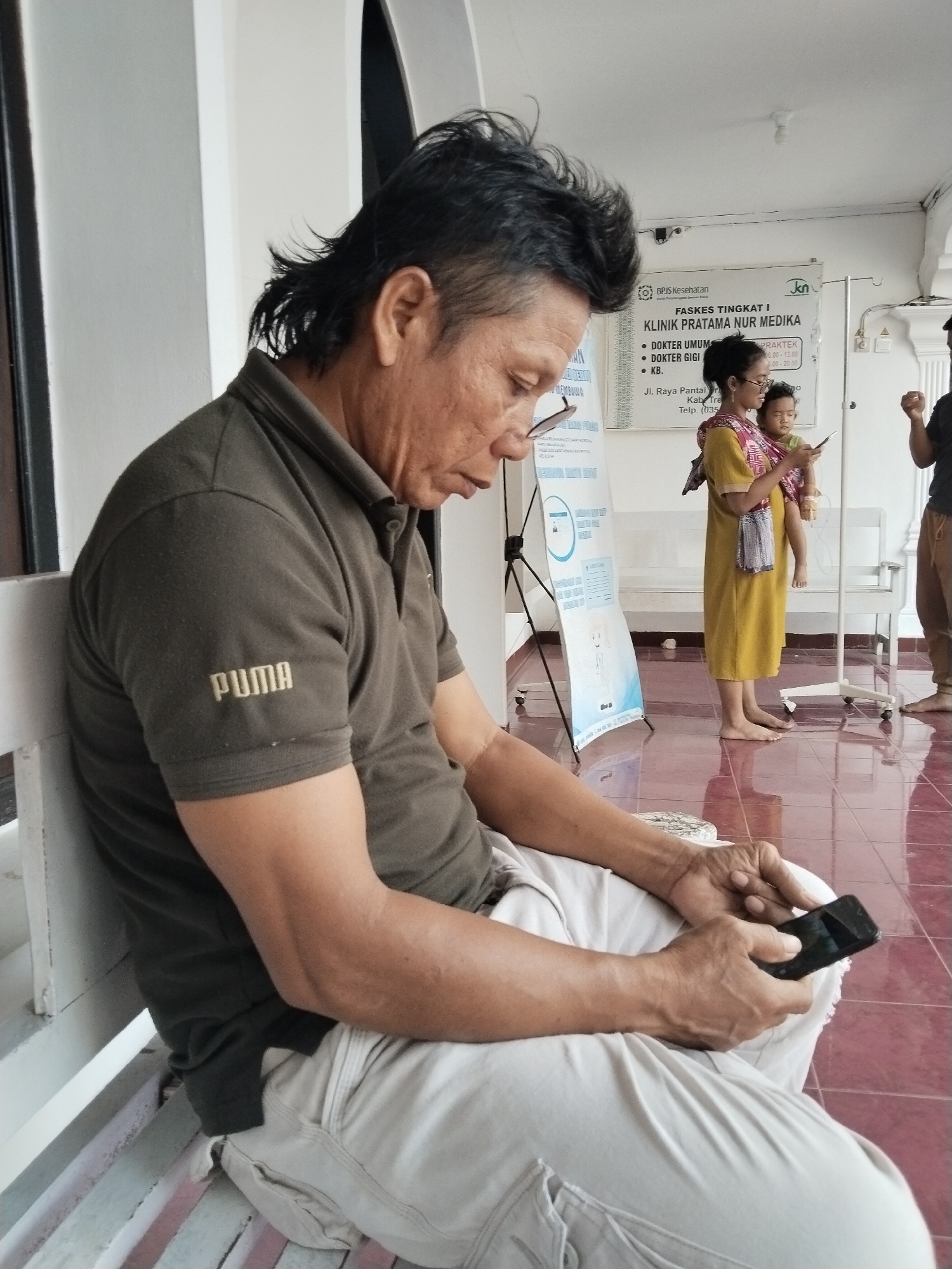 In Prigi, Indonesia, a man is seated on a white bench, looking down at his smartphone. He is wearing a dark olive green Puma polo shirt and light beige pants. His hair is styled in a mullet. To his right, a woman stands holding a baby wrapped in a colorful cloth sling, also looking at her phone. In the background, a white poster with text and graphics is displayed on a stand, and medical equipment like an IV pole is visible. The floor is tiled with a reddish-brown color, and a white pillar is present on the left side of the frame.