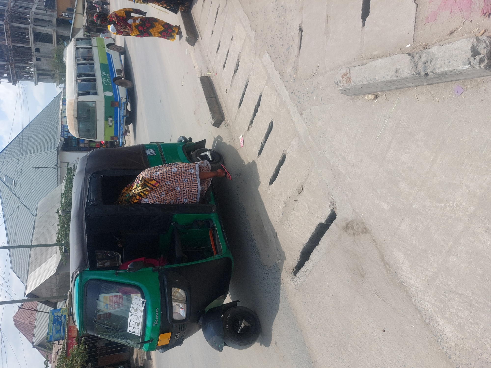 The image shows a three-wheeled vehicle parked at the side of a road in an urban area. The vehicle is green and black, and it appears to be a type of auto rickshaw, commonly known as a "bajaj" or "tuk-tuk" in many parts of the world. A person dressed in a patterned cloth appears to be exiting the vehicle or bending inside. A white and blue bus is visible in the background on the road, and there are pedestrians walking on the sidewalk. Buildings of varying heights are visible in the background. The street appears to have a concrete surface with a drainage system along the side. The location is most likely in Dar es-Salaam, Tanzania, as that is the city mentioned in the prompt.