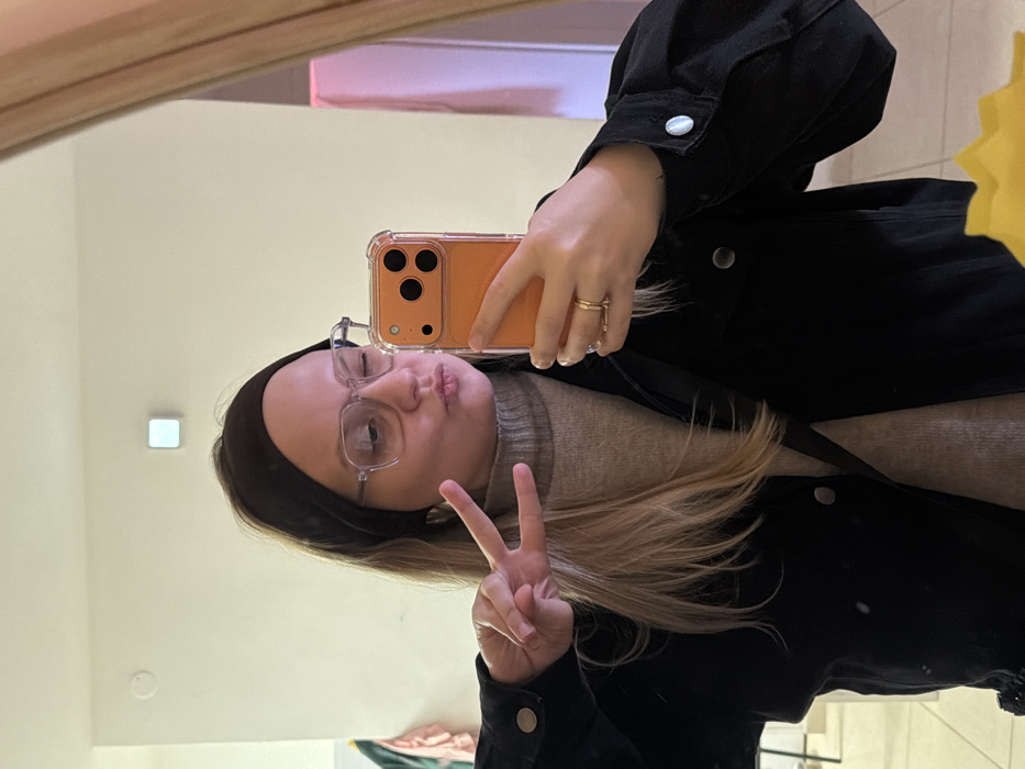 A female individual is captured in a mirror selfie, depicted from the chest up within an indoor setting. She has light-colored hair, partially covered by a dark headband, and wears clear-framed eyeglasses. Her attire includes a light brown turtleneck sweater and a black jacket with silver snap buttons. Her left hand is raised, forming a peace sign. Her right hand holds an orange-cased smartphone with a clear bumper, positioned to capture the reflection. Her facial expression features pursed lips and a slight squint of her right eye. A gold-colored ring is visible on the ring finger of her left hand. A black strap, consistent with a crossbody bag, runs diagonally across her chest. The background shows a plain, light-colored wall with a square, illuminated light fixture positioned centrally in the upper third. Blurred elements, possibly hanging clothing or furniture, are visible on the far left. A portion of a yellow object is present at the bottom right. The specific location within Tel-Aviv, Israel, cannot be visually determined from the image.