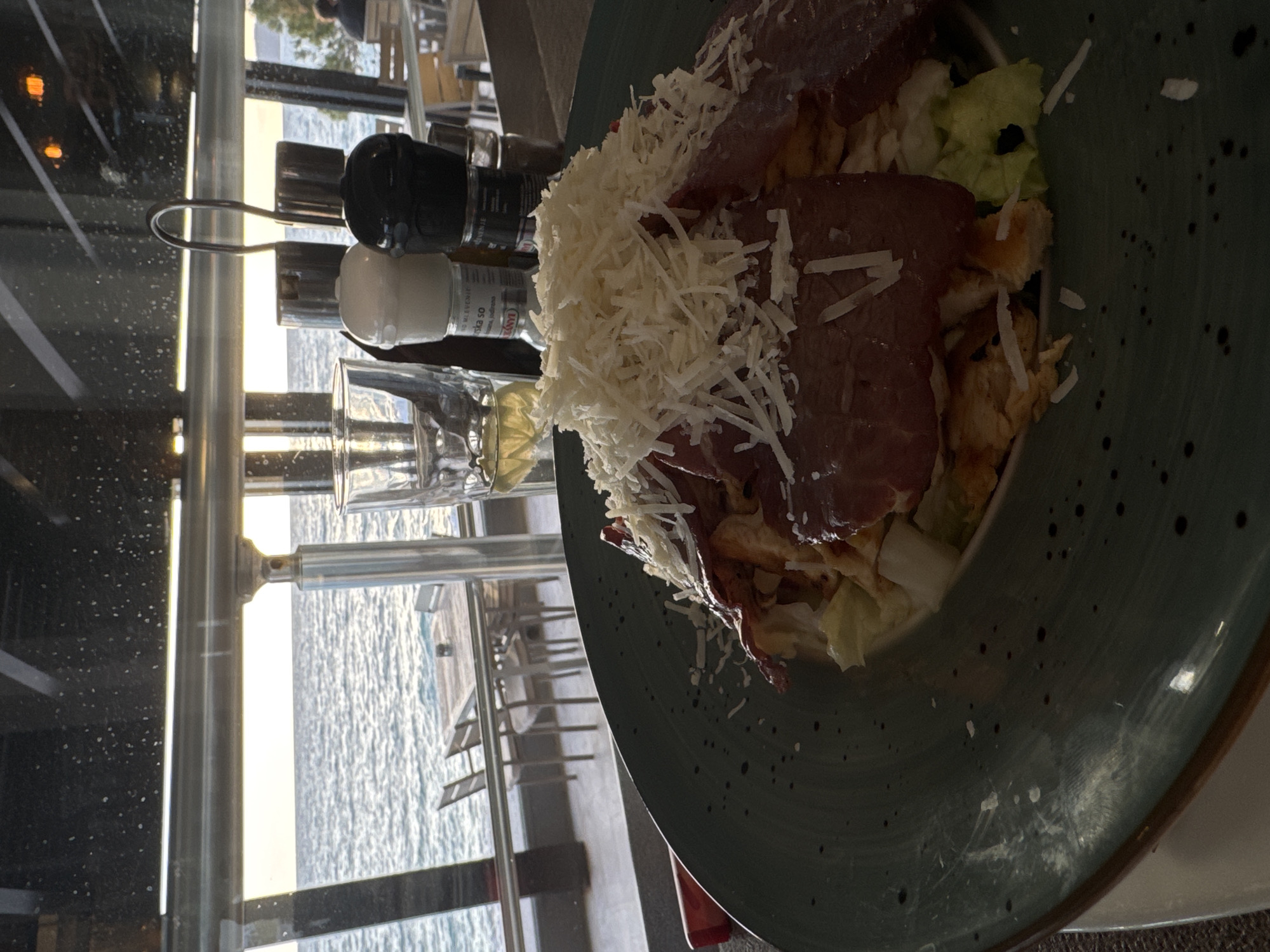 A close-up, tilted-angle shot shows a plate of food beside a window overlooking water, possibly the Adriatic Sea, indicative of a coastal location like Bar, Montenegro. The plate contains a salad with leafy greens, meat slices, croutons, and a generous topping of shredded cheese. A set of salt and pepper shakers and a drinking glass are positioned next to the plate, closer to the window. Through the window, a seascape is visible with a view of the water and, potentially, some structures on or near the water, suggesting a restaurant or dining area situated with a view. The environment implies a serene dining setting by the sea.