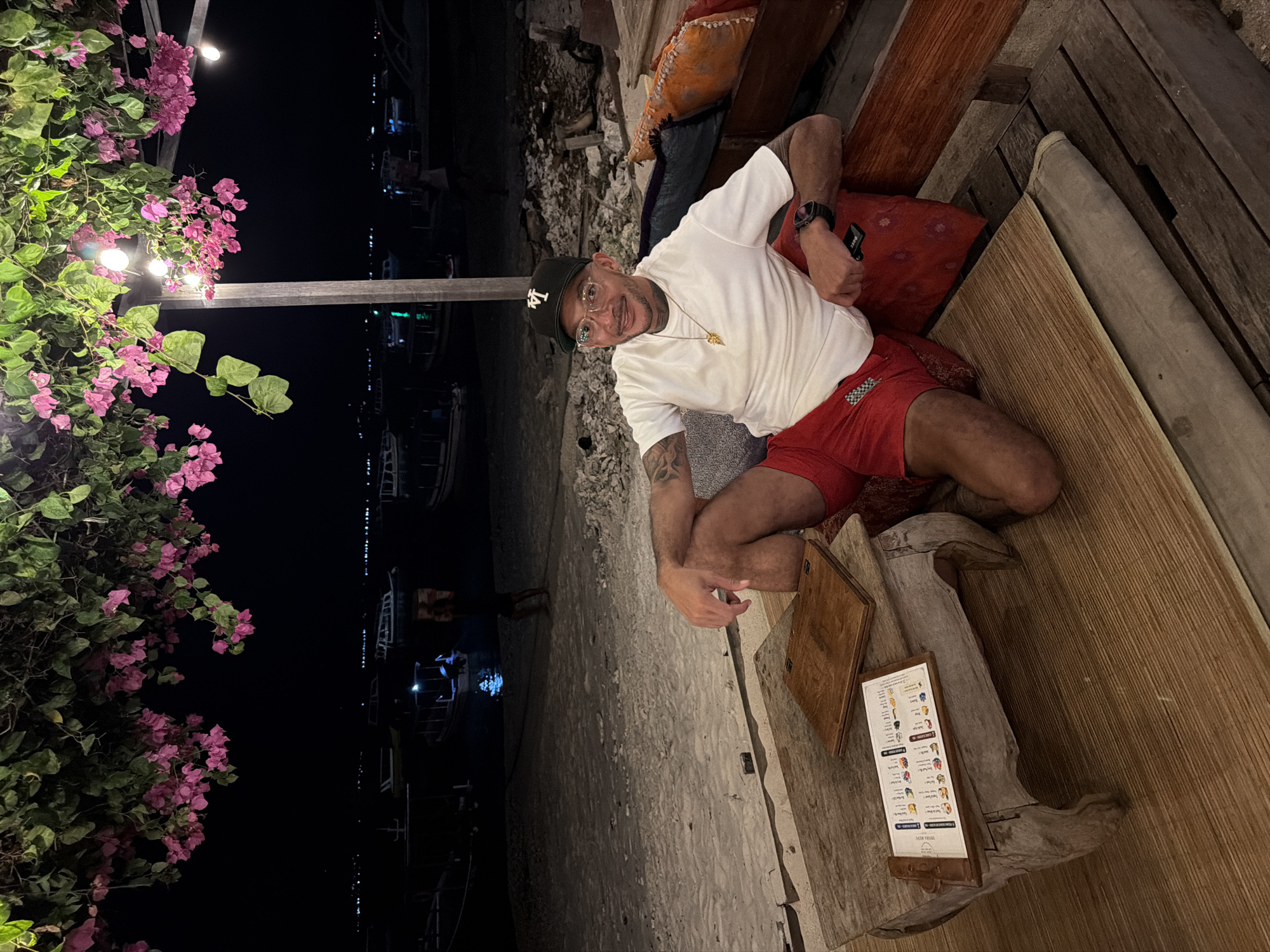 In a low-angle medium shot, a person is seated at a table on a beach at night. The person, wearing a white t-shirt, red shorts, and a baseball cap, is smiling at the camera. The table is made of weathered wood and has a menu and a wooden board on it. The person is sitting on a cushion on a wooden bench. In the background, there are several boats on the water and pink flowers hanging from an overhead structure. The scene appears to be in an outdoor restaurant or bar by the sea. The location is unknown, but the presence of boats and a beach suggests it could be a coastal area in Indonesia, such as Bali or Lombok.