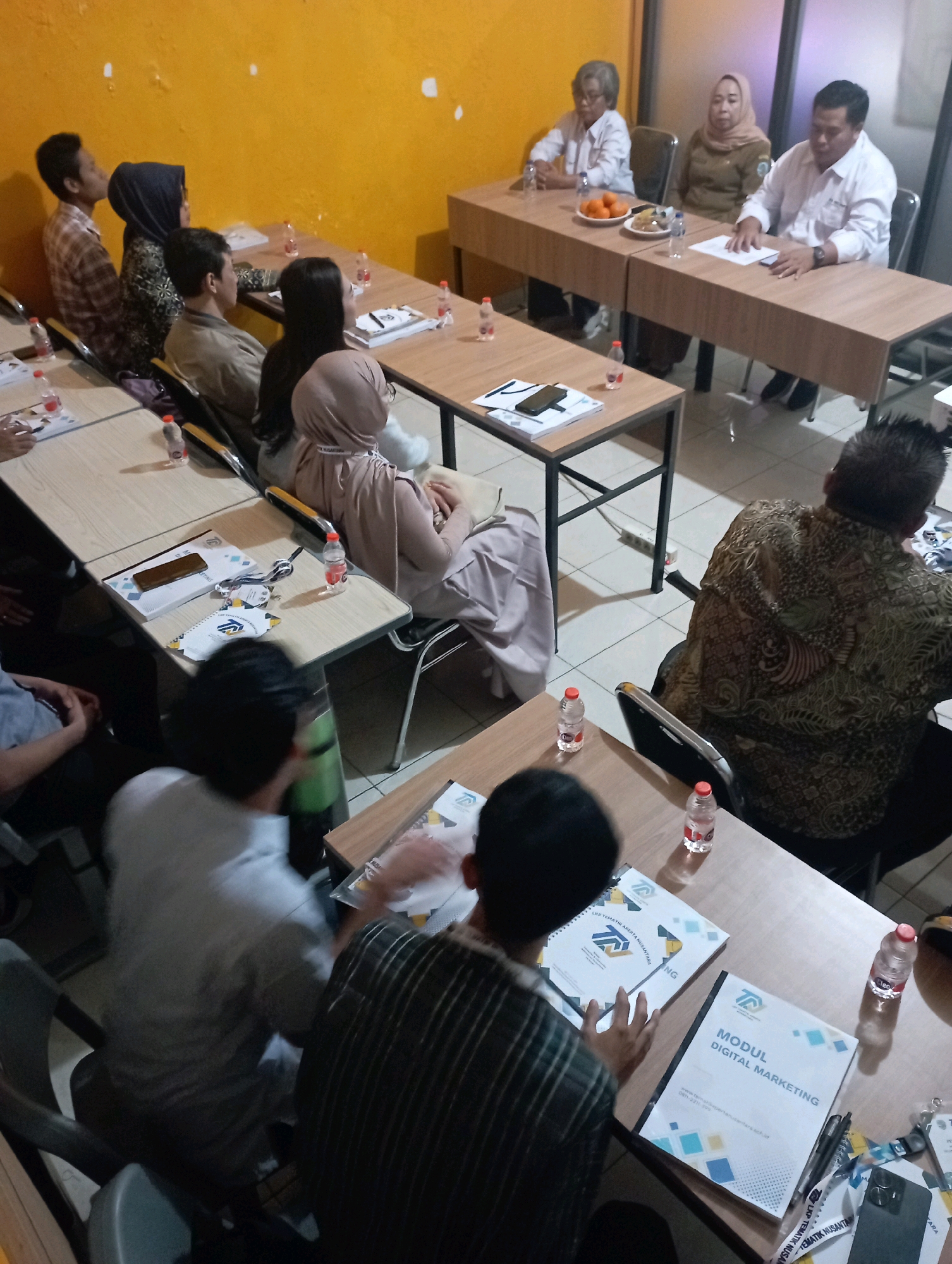 The image depicts an indoor meeting or seminar taking place in a room with yellow walls. Several individuals are seated at tables, some are facing forward, while others are engaged in discussion or looking at documents.
In the foreground, three individuals are seated, with two of them facing away from the camera. One person, in the center, is wearing a white shirt, and another, to the right, is wearing a black and white striped shirt. To the left, a person in a light-colored shirt is partially visible. On the tables in front of them, there are notebooks, pens, and water bottles.
In the mid-ground, several individuals are seated at tables arranged in a semi-circle. Some are wearing formal attire, such as white shirts. There are also water bottles and documents on the tables. One person is seen wearing a hijab and a light-colored dress, and another person is wearing a patterned shirt.
In the background, three individuals are seated at a long table. One person is wearing a white shirt and appears to be speaking. Another person, also in a white shirt, is seated next to them. A third person, wearing a hijab, is seated between them. On their table, there are plates of fruit, water bottles, and documents.
The overall scene suggests a professional or educational gathering. The presence of multiple tables, chairs, and presentation materials indicates a structured event. The yellow walls and the lighting suggest the room might be a conference room or a classroom. While the exact location is not specified in the image itself, the mention of "Tasikmalaya, Indonesia" in the prompt indicates the general geographical context of the image. The room appears to be moderately lit, with some artificial light sources contributing to the illumination. The flooring seems to be tiled.