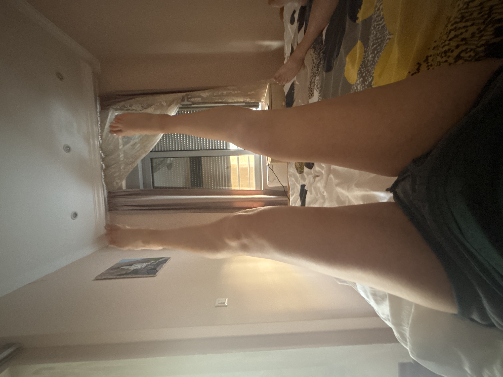 A person is lying in bed in a room in Budva, Montenegro. The bed has a patterned comforter and white sheets. The person is wearing dark shorts. A window with curtains is visible. There is a picture hanging on the wall.