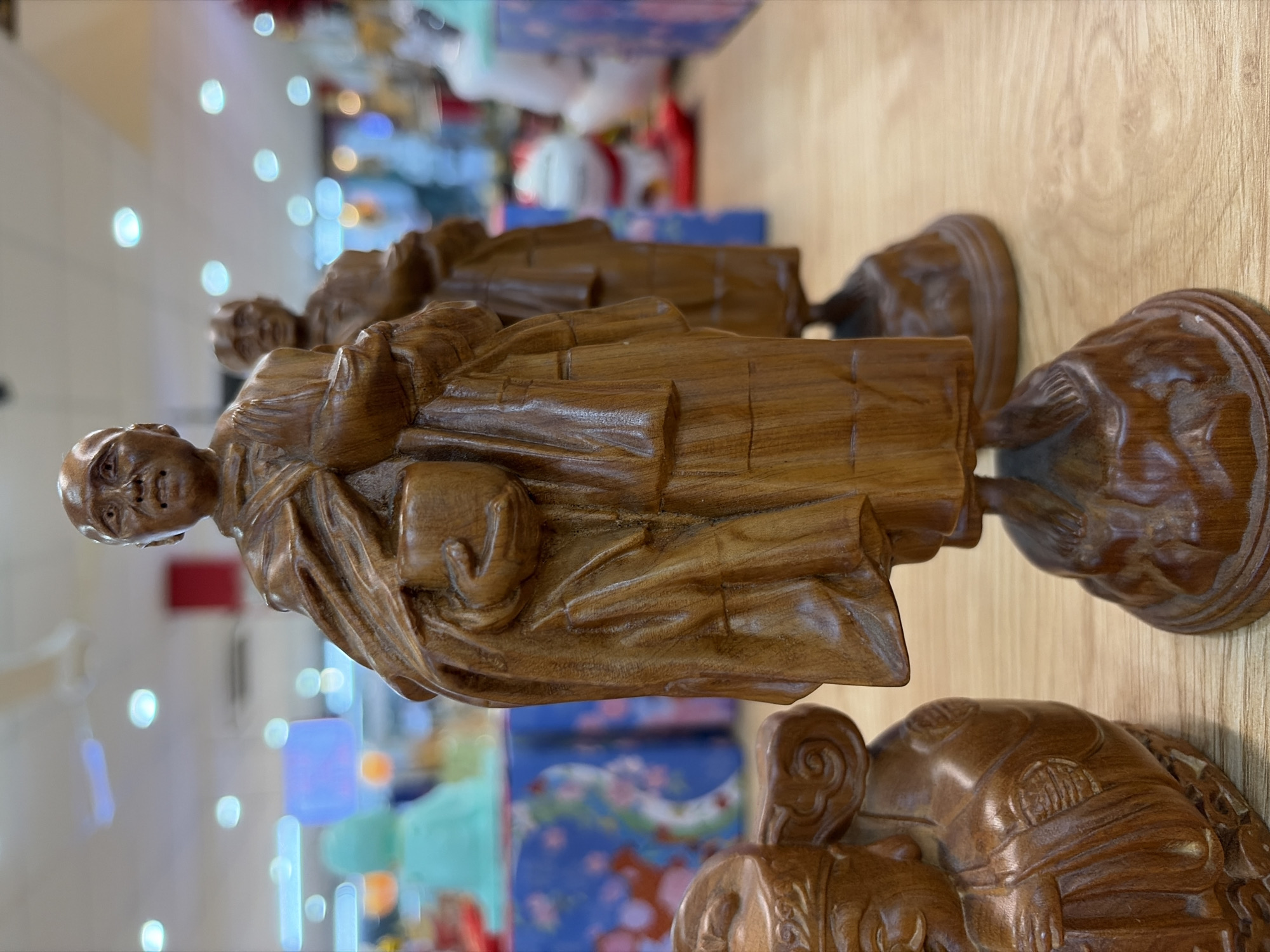 The image displays a close-up, eye-level shot of several wooden figurines on a wooden surface. In the foreground, a portion of a seated wooden figurine is visible on the left, featuring intricate carvings. To the right of this, a taller, standing wooden figurine of a bald individual wearing robes and holding a cup is prominently featured, with another similar figurine slightly blurred in the background. The background shows a retail environment with shelves and displays, including a blue and pink floral patterned cloth and other objects, suggesting the location is a shop in Đà Nẵng, Vietnam. The lighting is bright, with reflections visible on the surfaces.