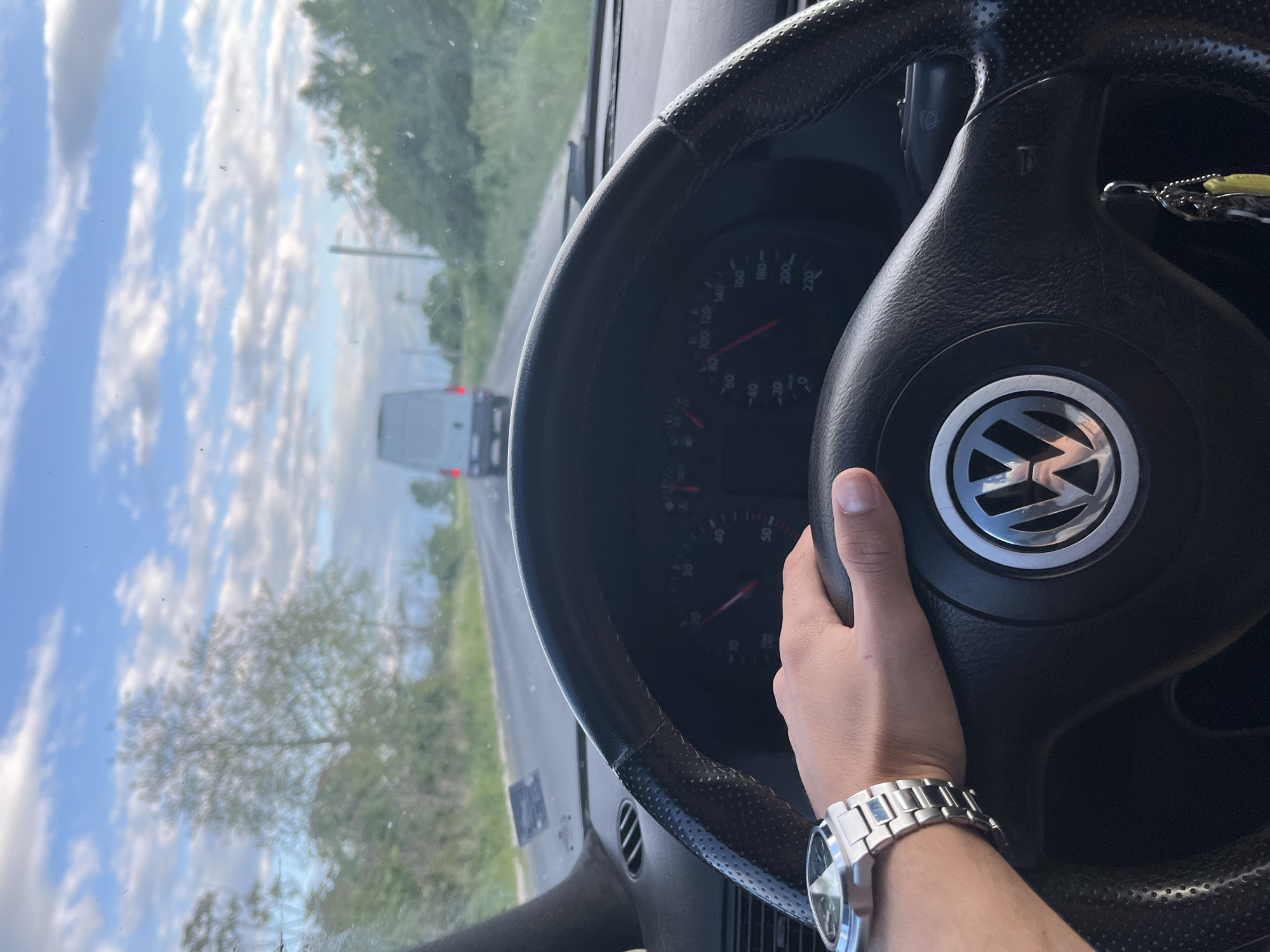 A person is driving a Volkswagen, based on the logo on the steering wheel. The vehicle is on a road, and a white van is visible in the distance ahead. The landscape includes trees and a cloudy sky. The location is within the vicinity of Dalgo Pole, Bulgaria, based on visual indicators. The person driving is wearing a silver wristwatch.