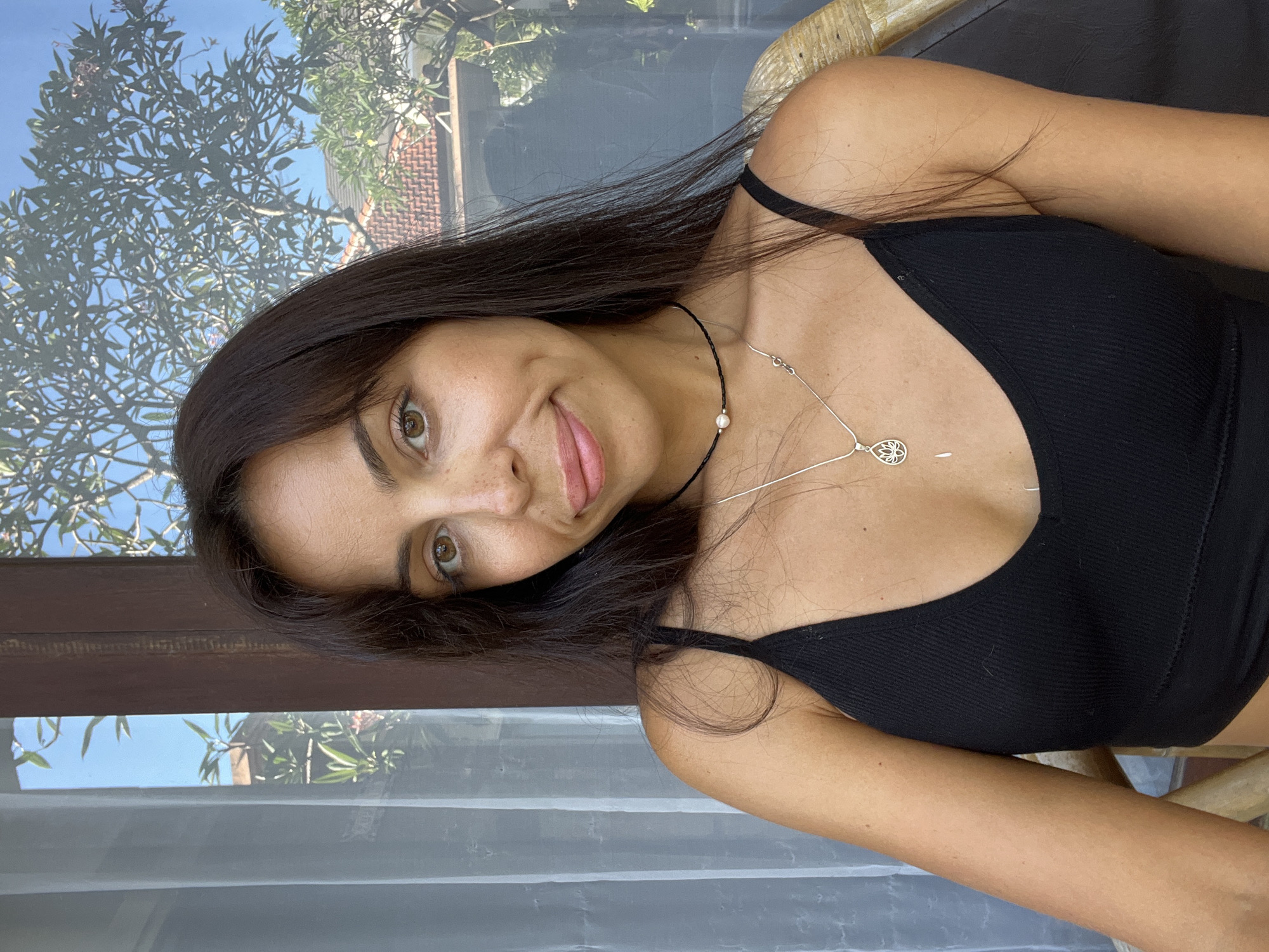 An image, rotated 90 degrees counter-clockwise, portrays a young woman with long, dark hair and striking brown eyes looking directly at the viewer with a gentle, slightly playful smile. Her head is tilted, and her tongue is subtly visible between her lips. She is wearing a black ribbed tank top and two necklaces: one is a black cord adorned with a single white pearl, and the other is a silver chain featuring a delicate, circular pendant, possibly a stylized lotus flower.
The woman is seated indoors, positioned near a window that reveals a clear view of a sunny outdoor environment. The background displays a vibrant blue sky, dense green foliage of trees, some featuring pink or purple blossoms, and parts of a distinctive reddish-brown tiled roof from an adjacent building. A dark wooden window frame and a sheer white curtain are also visible, framing the outdoor scene. The bright natural light indicates it is daytime with clear weather. The tropical vegetation and architectural cues are consistent with the location context of Kuta Selatan, Indonesia. No text is visible in the image.