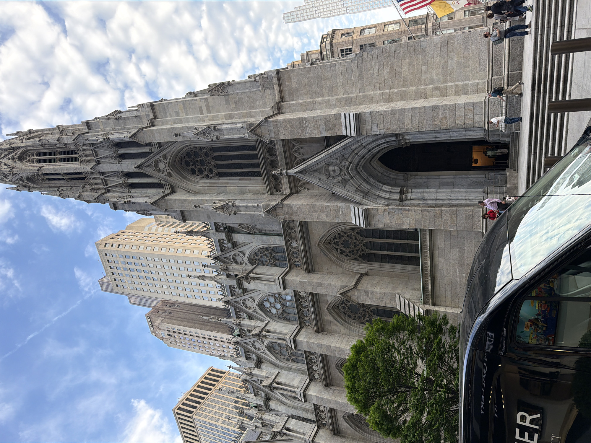 This photo, taken at a low angle, captures Trinity Church in the Financial District of New York City. The church, a Gothic Revival structure, occupies the left side of the frame. It is built of grey stone and characterized by pointed arches, ornate tracery, and a tall steeple reaching towards the sky. Buildings of varying heights are adjacent to the church. Several people are near the front entrance of the church; others are on the stairs. The sky, a mix of blue and white, contains scattered clouds. The right side of the frame is partially occupied by a black vehicle.