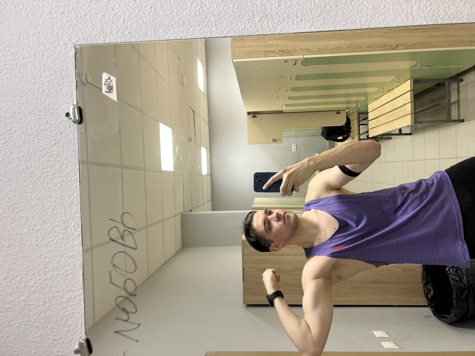 A man stands in front of a mirror in what appears to be a locker room. He is flexing his biceps while holding a smartphone, presumably taking a selfie. He is wearing a purple tank top and dark-colored bottoms, with a black wristband on one arm. The reflection in the mirror shows locker room benches and walls with graffiti. The room is brightly lit with overhead fluorescent lighting.