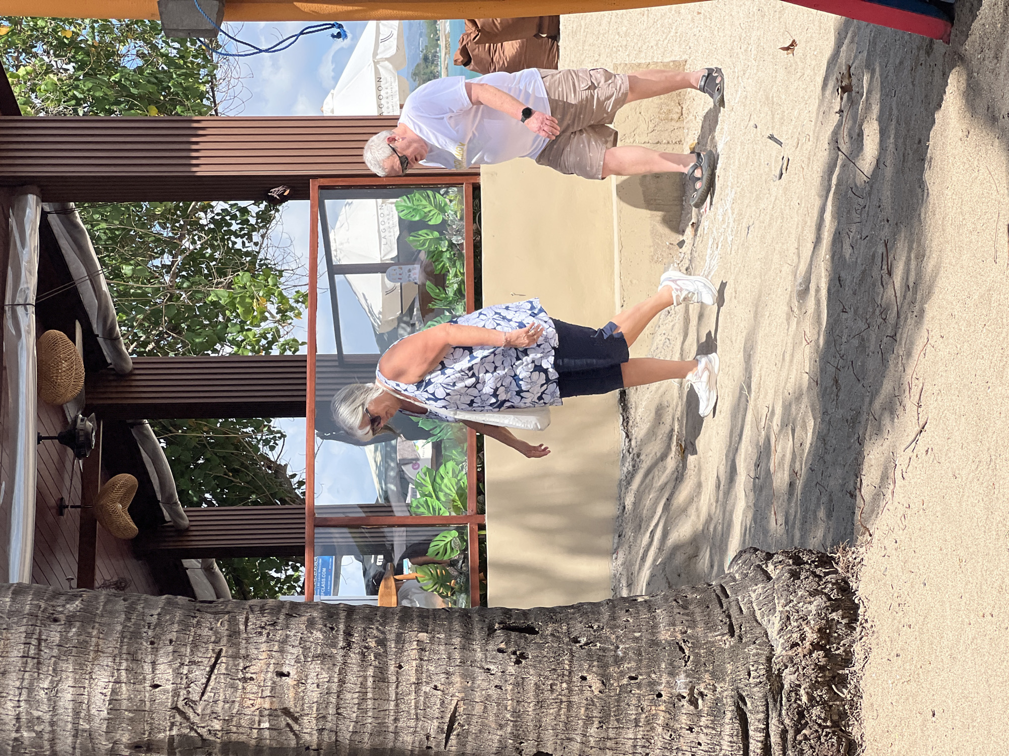 In Kuta, Indonesia, a sandy pathway adjacent to a building with wooden accents features two individuals walking. A large palm tree trunk dominates the left foreground. The woman on the left, wearing a white and blue floral sleeveless top and dark shorts, carries a white tote bag. She has short, gray hair and sunglasses. The man on the right wears a white t-shirt, patterned khaki shorts, sunglasses, and sandals. He has short, gray hair and a watch on his left wrist. They are walking in the same direction, with the woman slightly ahead of the man. Behind them, a building with glass windows displays tropical plants. In the background, white umbrellas are visible, suggesting an outdoor seating area. The ground is sandy with scattered small debris and shadows cast by the palm tree.
