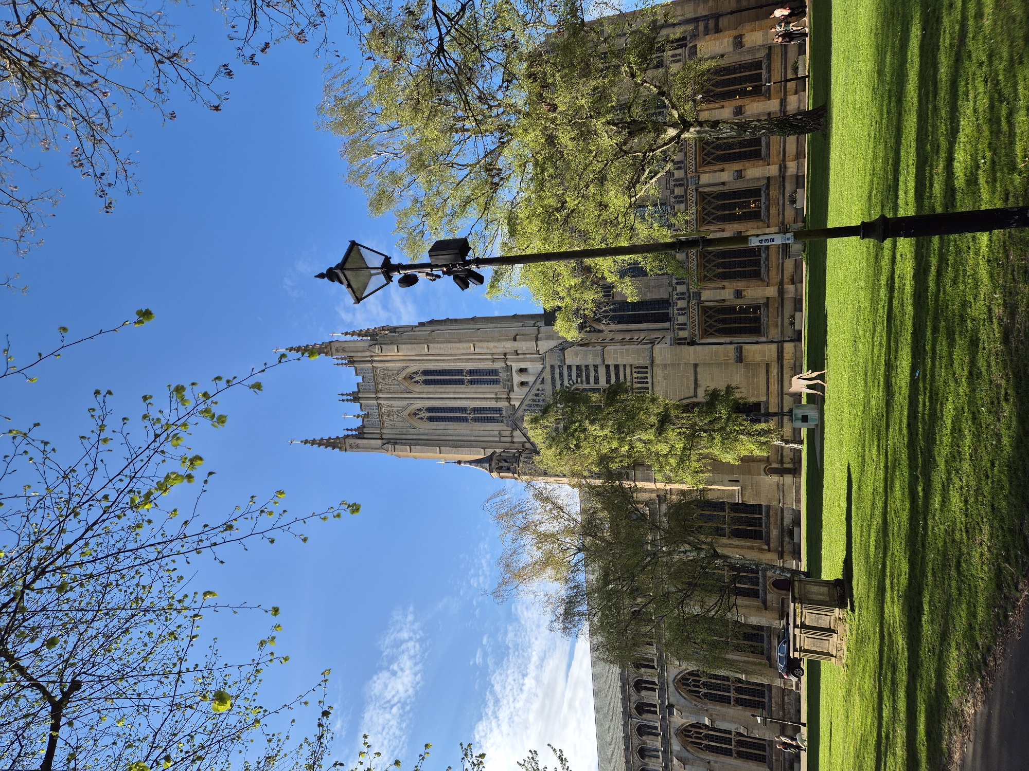 The image shows a tall, ornate cathedral in Bury St Edmunds, West Suffolk, United Kingdom, viewed from a low angle. The cathedral is made of light-colored stone and features multiple pointed spires, arched windows, and intricate architectural details. A black lamppost is centrally placed in the foreground. The cathedral is partially obscured by trees with budding green leaves. A stretch of green lawn is to the right of the building, casting long shadows from the trees. A few people are walking in front of the cathedral. The sky is blue with scattered white clouds.