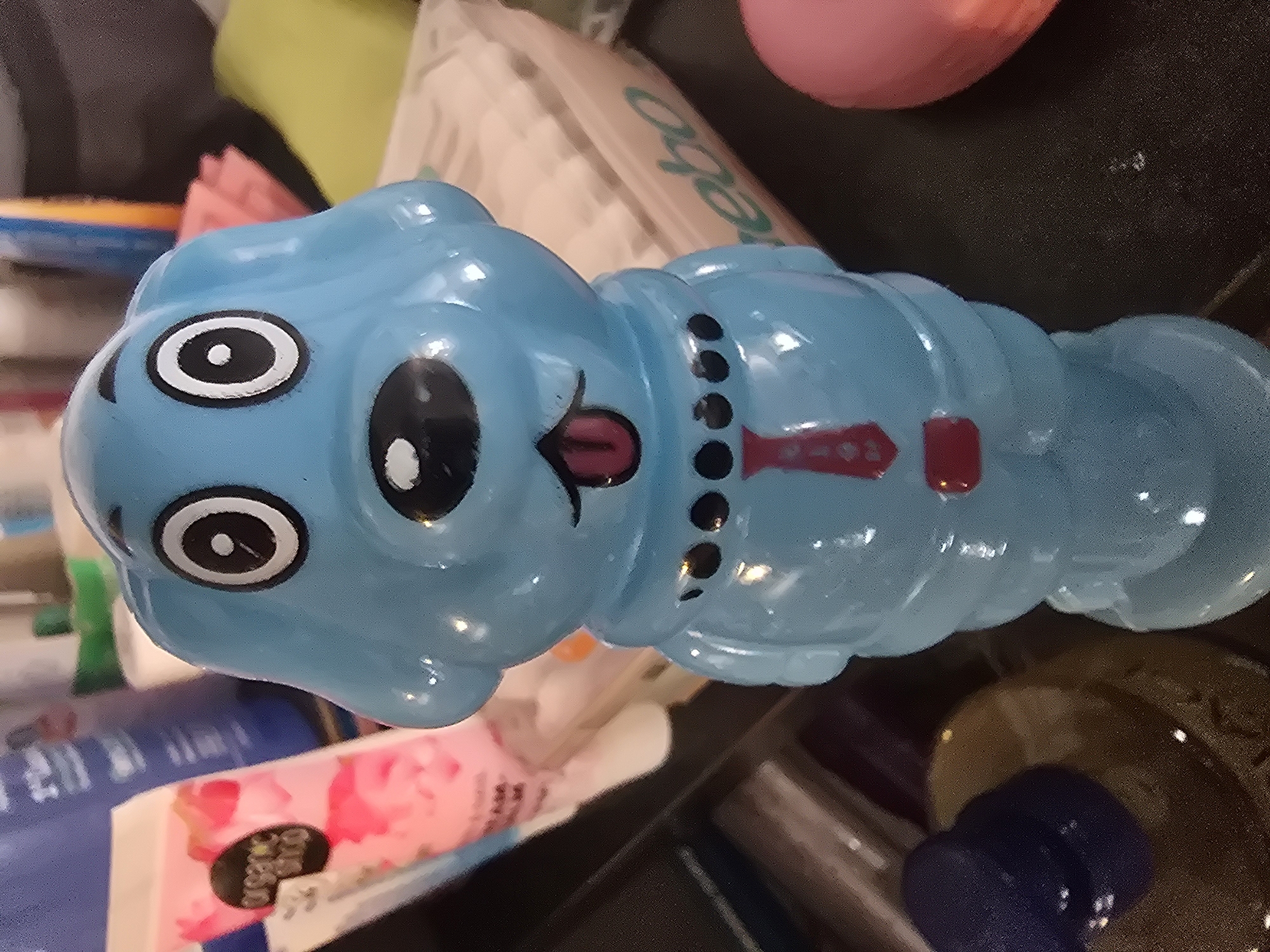 A close-up, eye-level shot shows a bright blue, plastic dog figurine with black eyes and a pink tongue sticking out. The figurine is wearing a red tie with a white pattern and a black collar with three black dots. In the background, out of focus, are various toiletries including a pink tube labeled "organic shop," a blue bottle, and a yellow object. The figurine is positioned in the foreground on a dark, reflective surface. The location is Unknown, Unknown.