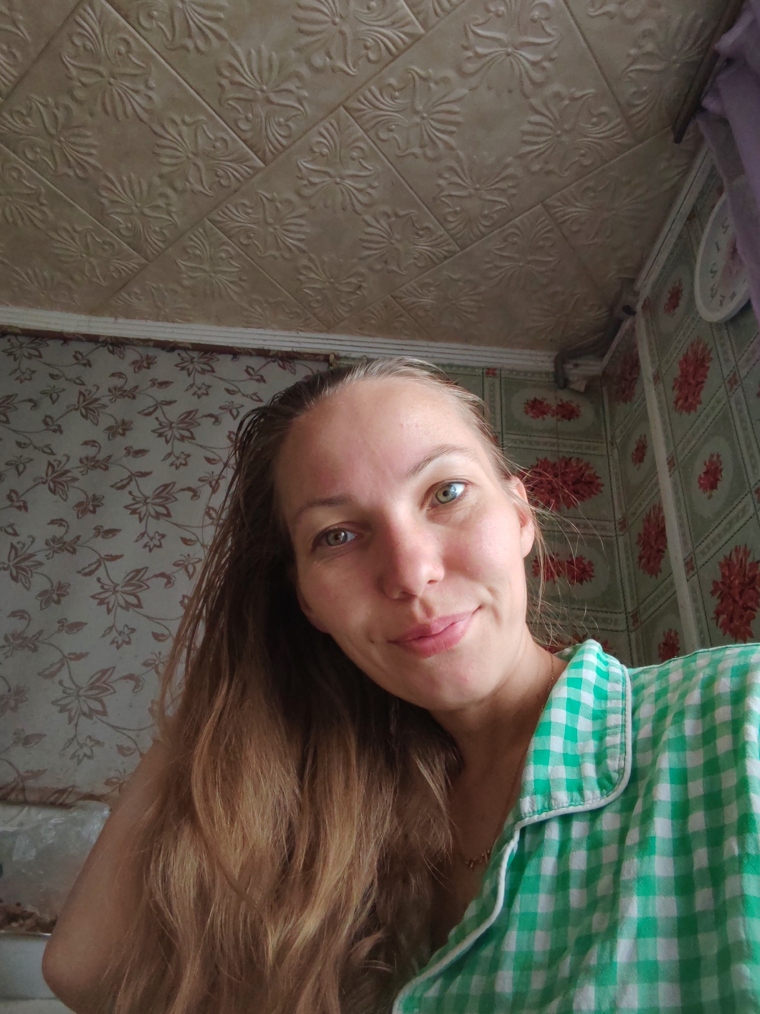 A woman is depicted indoors in what appears to be a residential space, with a low-angle view of her face. She has fair skin, hazel eyes, and long brown hair. She's wearing a green and white checkered shirt. The background includes patterned wallpaper with floral designs and a textured ceiling. The location is reported as Чухлэм, Russia, though this is not evident from the image. There are no apparent actions or interactions occurring in the image.