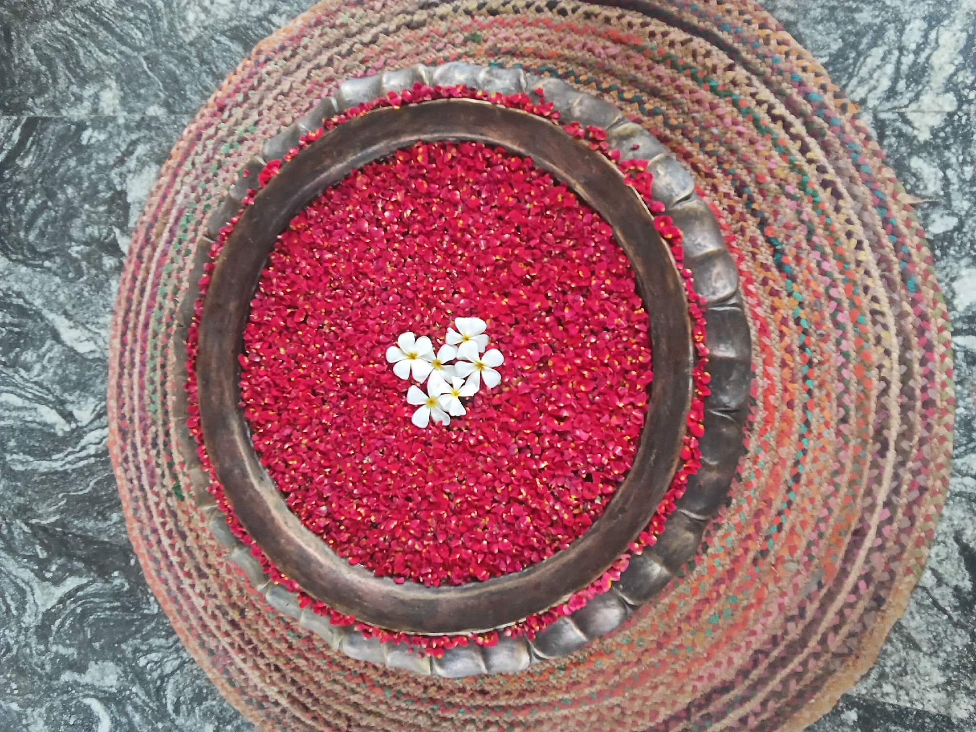 A top-down shot presents a circular metal bowl filled with red flower petals, with five white flowers clustered in the center. The bowl sits atop a round, woven rug with multicolored strands. The background consists of a gray, mottled surface. The image was taken in Ashvem, India.