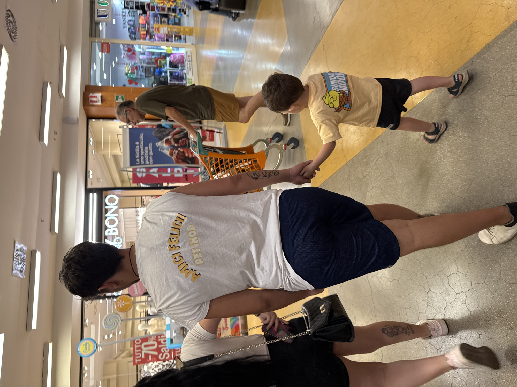 The image depicts a scene inside a commercial establishment, likely a shopping mall, in Palermo, Italy. In the foreground, a young boy in a yellow t-shirt and black shorts is walking hand-in-hand with an adult male wearing a white t-shirt and dark shorts. The t-shirt of the adult male has text that reads "AMICI FELICI PET SHOP" in yellow lettering. Behind them, a woman with long dark hair, wearing a black top and shorts, is also walking, carrying a black handbag. Another adult male, wearing glasses, a green t-shirt, and brown shorts, is visible in the middle ground, pushing an orange shopping cart. The environment appears to be a bright, well-lit indoor space with tiled flooring in yellow and grey. Storefronts with signs and merchandise are visible in the background, including one with the text "BONO" and another with "YANKEE". The overall scene captures a family or group of individuals navigating a shopping area.