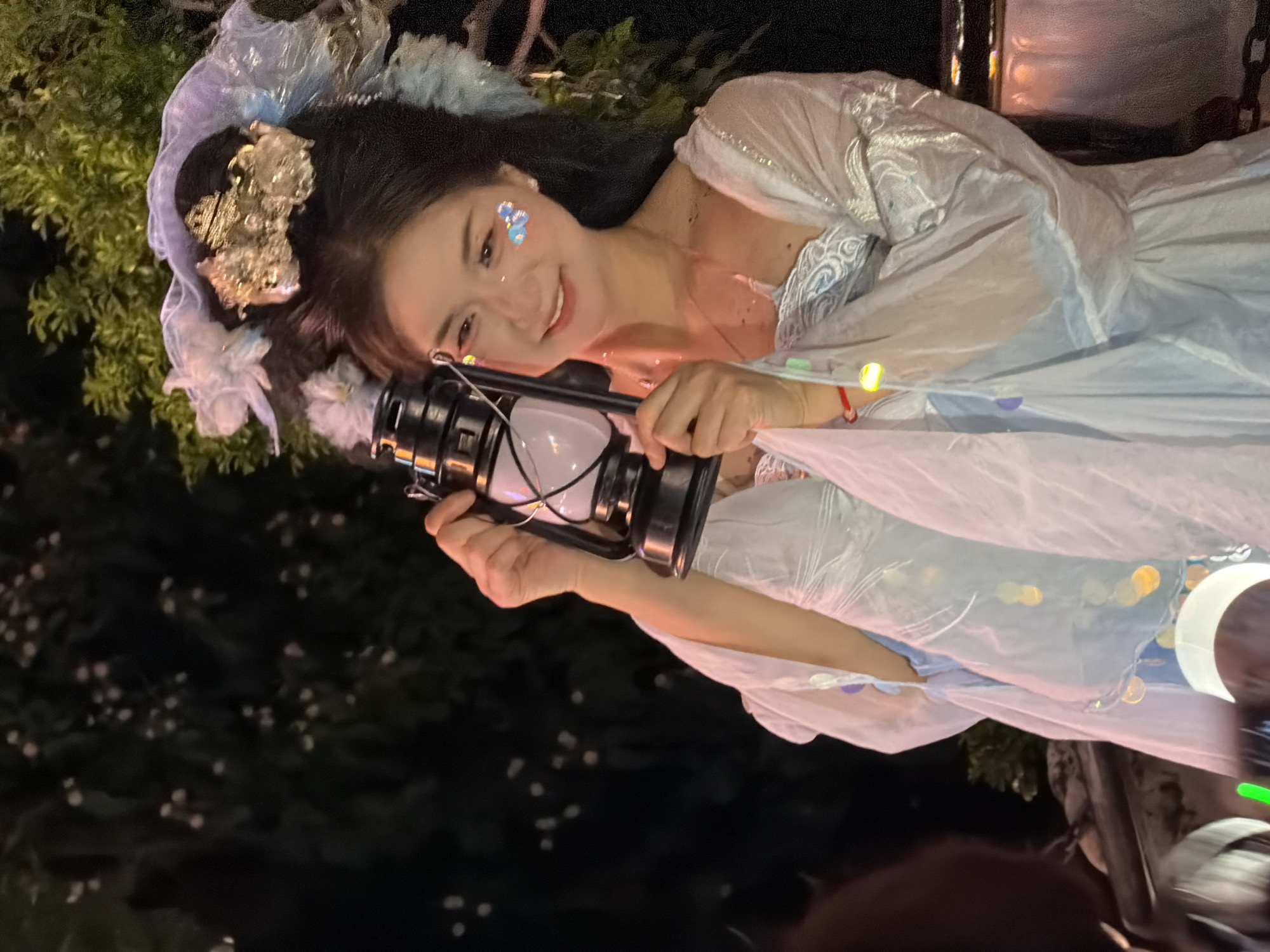 A woman, adorned in a light blue traditional Chinese dress and elaborate headwear, is captured in a medium shot as she holds up a black kerosene lantern. The woman's makeup includes glitter on her cheeks. She appears to be outdoors at night, with blurred foliage and lights in the background. The scene suggests a festive or cultural event, possibly taking place in Yuzhong District, China, given the implied cultural context of the attire and lantern. The lighting is dim, highlighting the woman and the lantern she holds.