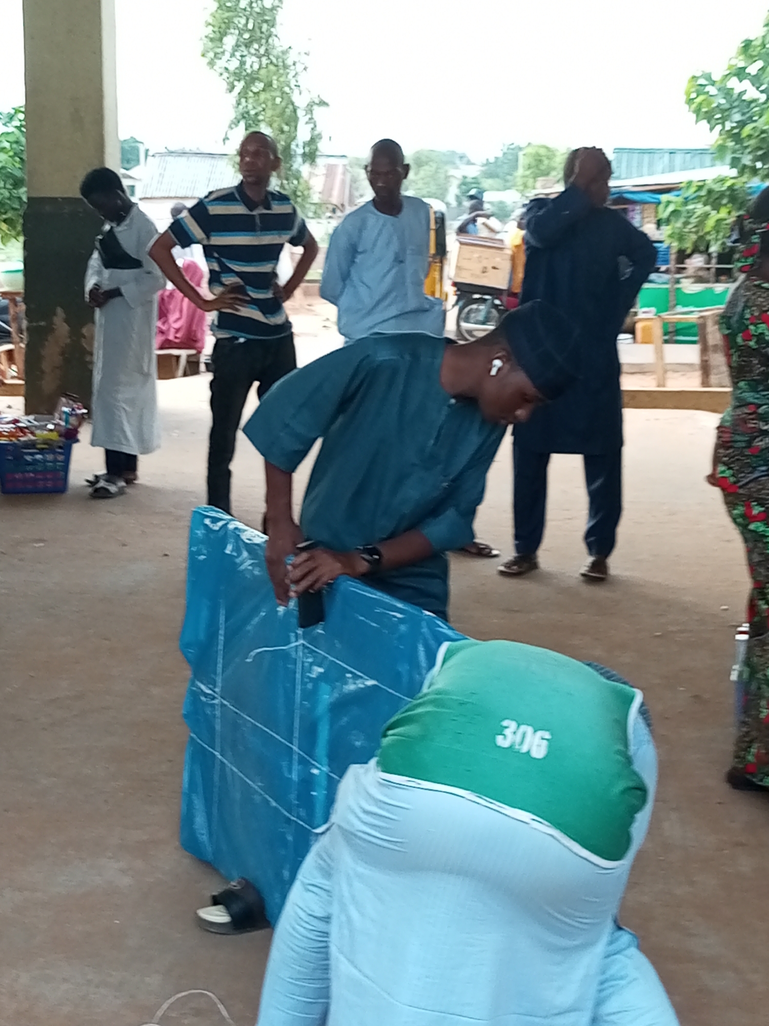In Gombe, Nigeria, a group of individuals are gathered outdoors, with a partially visible structure suggesting a market or an open-air venue. In the foreground, a person in a teal tunic and a black cap, wearing white earbuds, is bending over a large, blue-wrapped object that is tied with white string. The person is holding a dark object, possibly a phone, in their right hand, and appears to be examining or manipulating the blue package. To the left, a person in a white robe stands with their head bowed, and to the right, a person in a dark blue tunic and cap is seen from the back, with their hands on their hips. Another individual in a striped shirt and black pants is standing with hands on hips in the background. A market stall with a basket of items is visible to the far left. The ground is a dusty, light brown surface. In the background, buildings with corrugated iron roofs and green foliage are present, indicating an outdoor, semi-urban or rural setting.