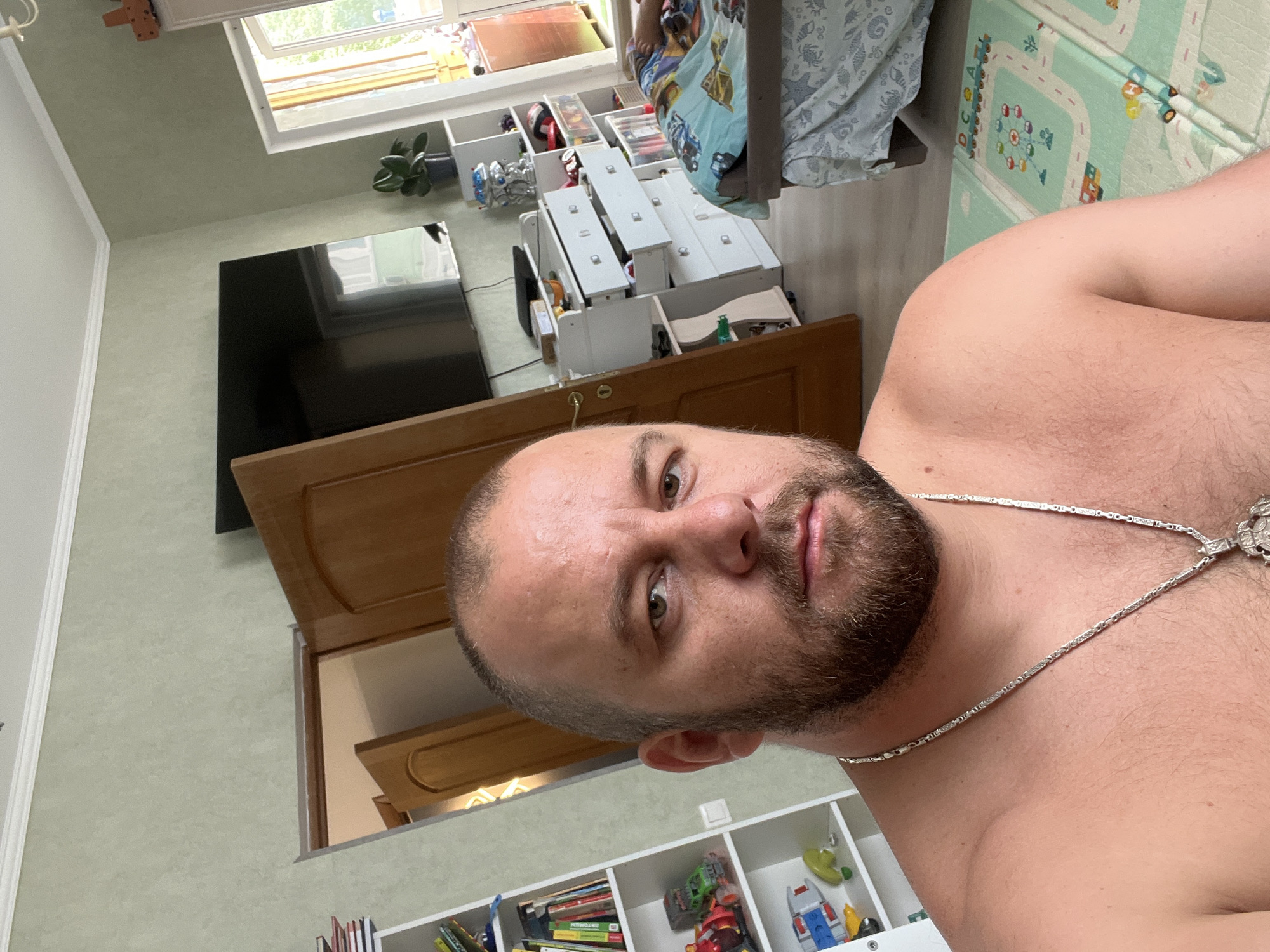 The image depicts a man in the foreground, taking a selfie in what appears to be a bedroom or living area. The man has a beard and is wearing a silver chain around his neck. He is shirtless.
In the background, there is a white bookshelf filled with books and toys. To the right of the bookshelf, there are several wooden doors, one of which is open, revealing another room. Further into the background, a television is mounted on the wall above a chest of drawers with some drawers open. A bed with a person lying on it and colorful bedding is visible on the far right of the image. A large window or door leading outside is also present on the right side of the room. The walls are painted a light green color. The floor appears to be laminate flooring, with a colorful play mat visible in the bottom right corner.
Based on the available information, it is not possible to determine the specific location in the city of Sochi, Russia.