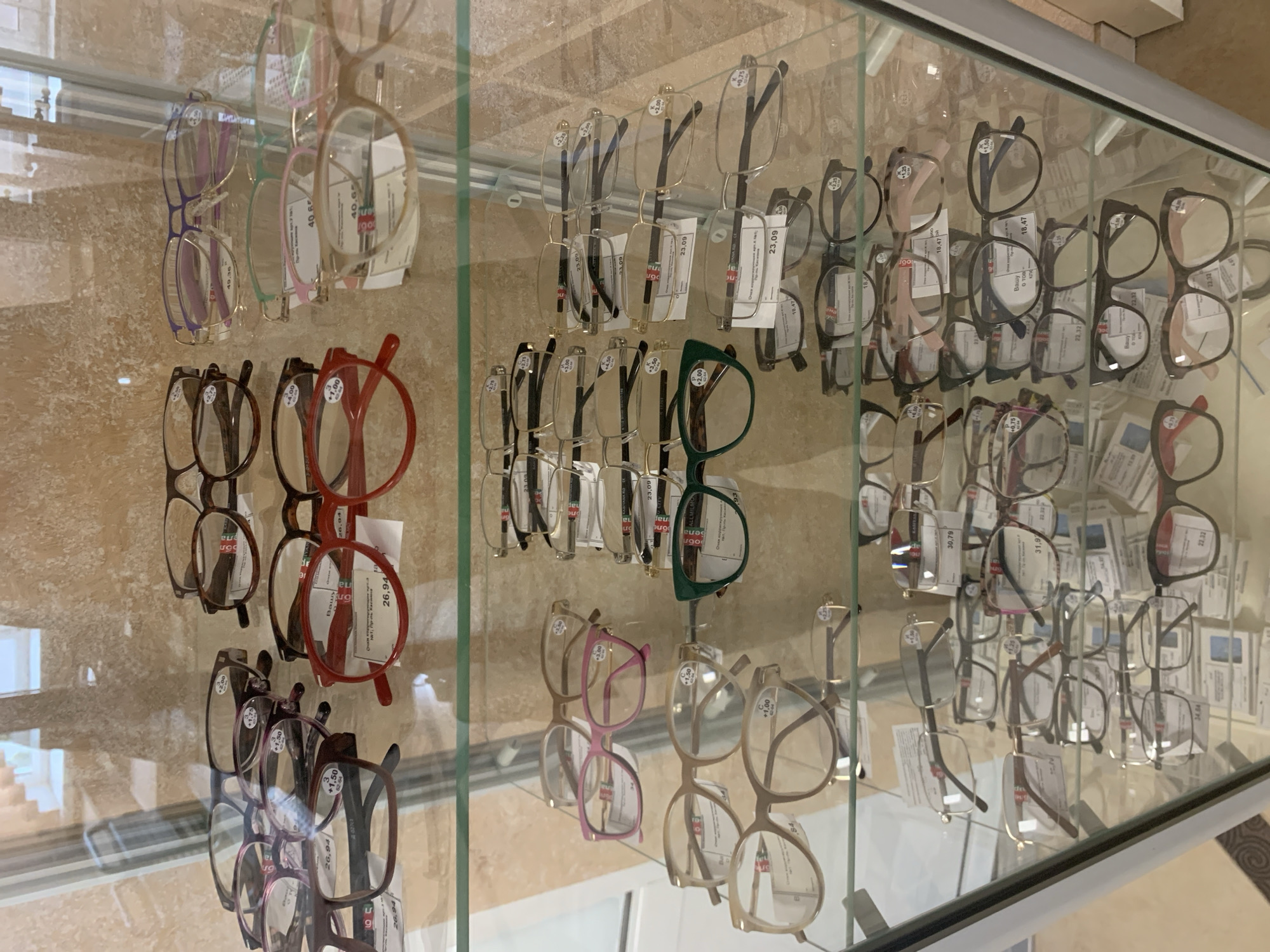 The image shows a display of reading glasses in a store. The glasses are arranged on multiple shelves behind a glass case. The shelves are organized with different styles and colors of frames. Some of the glasses have price tags and labels indicating their magnification. The store appears to be an optical shop or a department store with an optical section. The environment suggests an indoor retail setting. There are no people visible in the image.