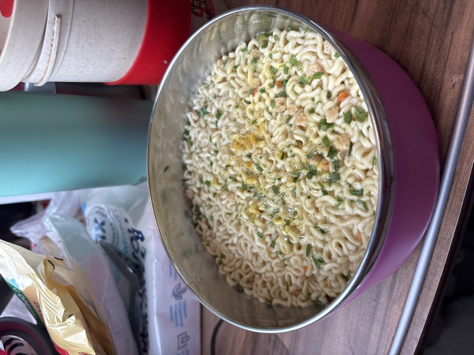 This image displays an indoor scene in the city of Tatarsk, Russia. The central focus is a round, stainless steel bowl with a glossy pink base, resting on a brown, wood-grain surface. The bowl contains instant noodles, which are partially submerged in liquid and topped with green, chive-like pieces, yellow seasoning powder, and small, light-colored dehydrated vegetable or protein fragments. Small bubbles are visible on the surface of the liquid. In the upper left background, a crumpled yellow plastic bag is visible, partially obscuring a white plastic package marked with Russian text, including "TIX XL" and "РОДНОЙ". To the upper right, a light teal-colored cylindrical container, possibly a thermos, stands next to a stack of two beige, lidded containers, with the bottom container featuring a red silicone band. A thin, metallic strip runs horizontally across the lower portion of the wooden surface, beneath the bowl.