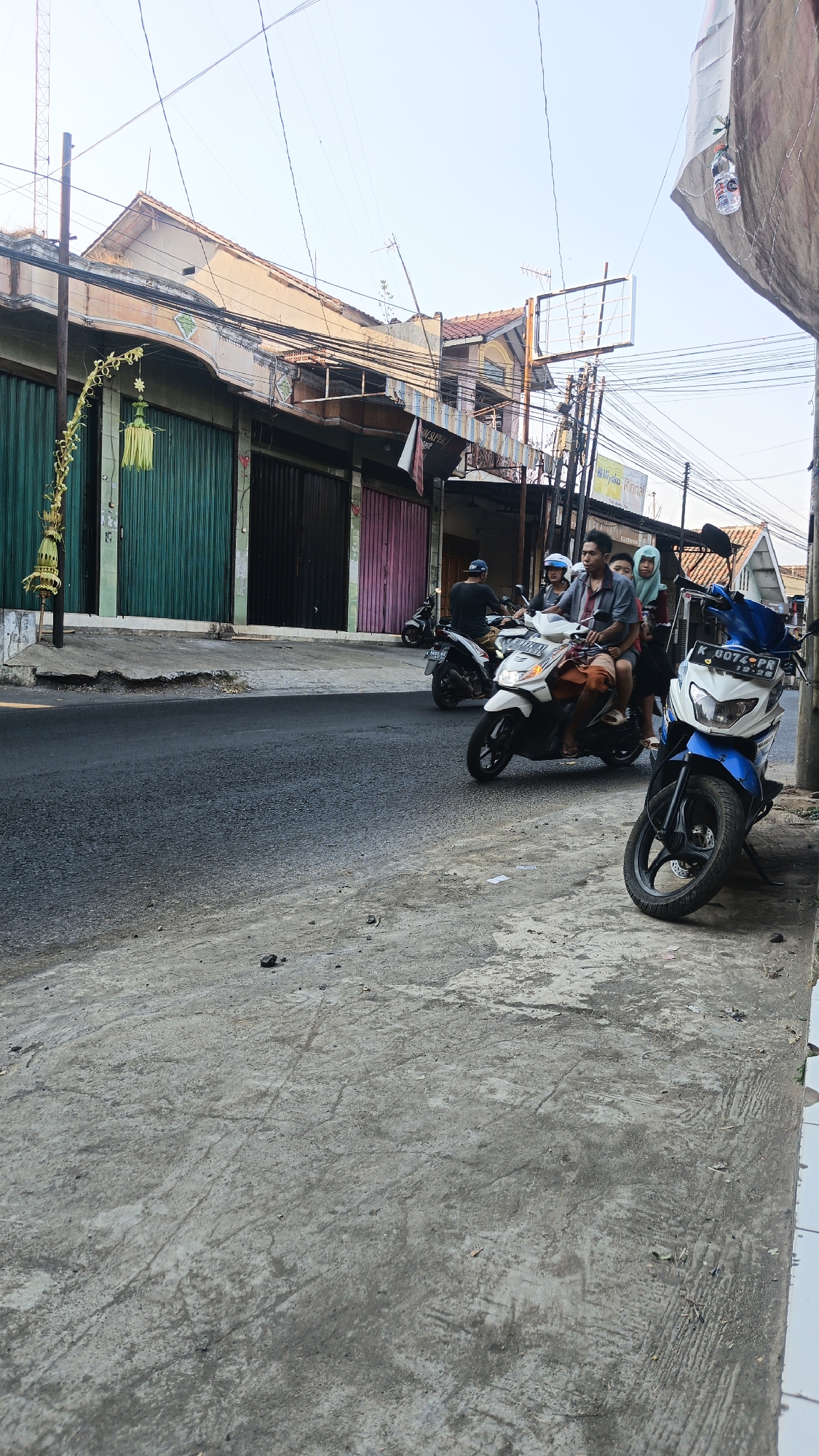 The image depicts a street scene in Dawe, Indonesia, with a focus on several motorbikes and people. In the foreground, a blue and white motorbike is parked facing left, with its license plate reading "K 6074 PR". To its right, a concrete sidewalk runs along the edge of the road. Further back, a white motorbike is carrying three passengers: a driver, a person in the middle, and a person at the rear wearing a light blue hijab. Another motorbike, a black and white model, is visible behind them with a single rider wearing a dark cap. The street is lined with shops, identifiable by their rolling metal doors, some in green, black, and pink. The buildings appear to be two stories high, with wires crisscrossing overhead. The overall environment suggests a typical urban setting in Indonesia, with a clear blue sky and warm daylight illuminating the scene.
