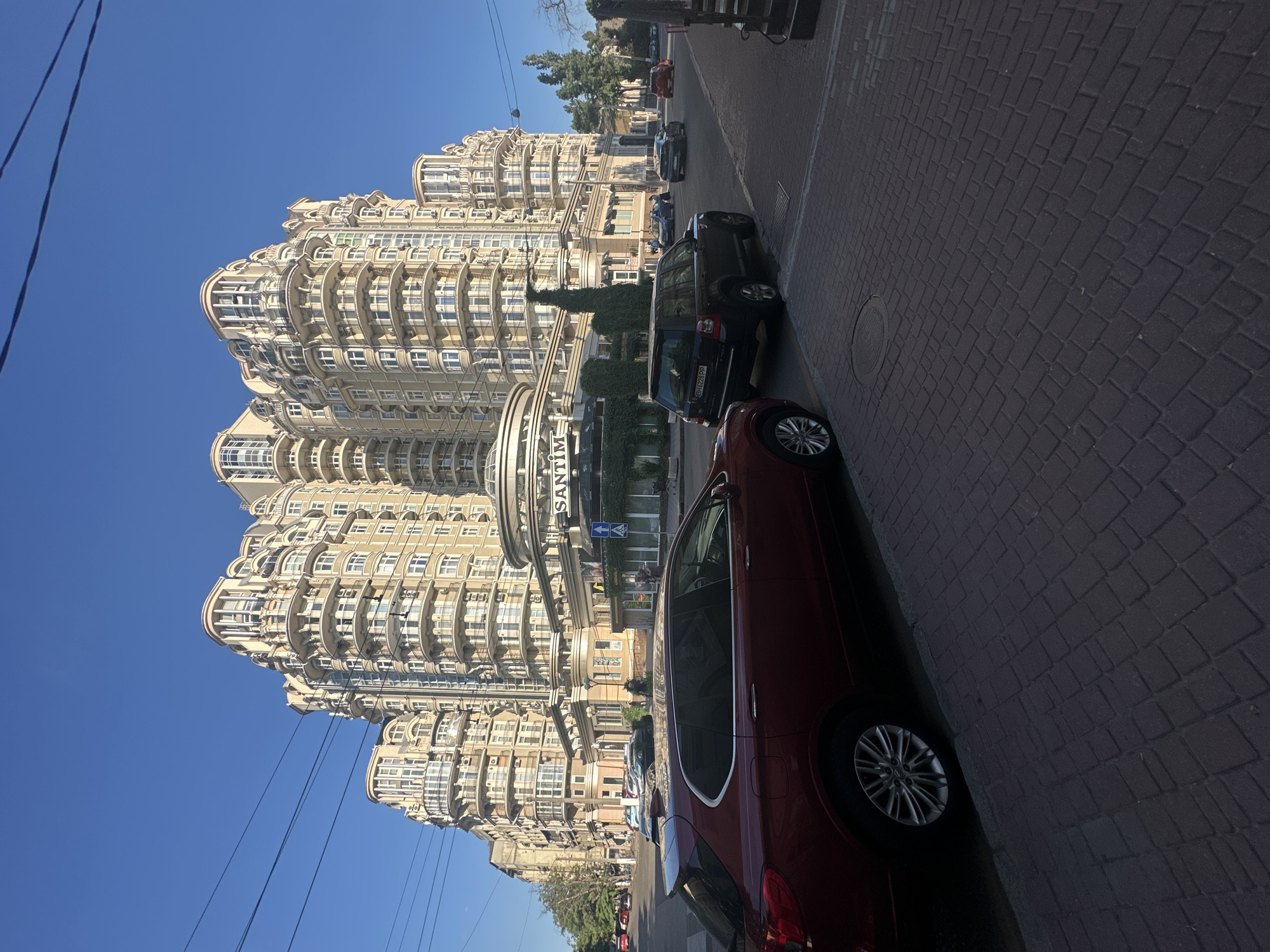 The image depicts a street scene in Odesa, Ukraine, characterized by a large, ornate apartment building with a pale facade and numerous curved balconies. Several cars are parked along the brick-paved street in front of the building, including a dark-colored SUV and a red sedan. The sky is a clear, vibrant blue, suggesting it is daytime with good weather.
The building's architecture is a prominent feature, with repeating rounded balconies creating a distinctive visual rhythm. Greenery is visible, with trees and bushes near the building's entrance and along the street. The presence of power lines suggests an urban environment.
The activity taking place is primarily passive observation of the urban landscape. The cars are parked, and no people are actively engaged in a discernible event. The overall mood is one of quiet urban life under a bright sky.
Notable details include the textured brickwork of the street and the clean, modern appearance of the cars. One of the buildings appears to have signage that reads "SANTIM." The time of day is clearly daytime due to the bright sunlight casting shadows and illuminating the scene. The clear blue sky indicates pleasant weather.