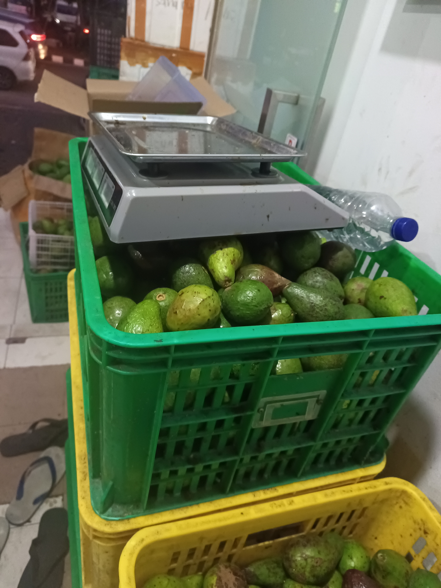 An outdoor market scene in North Jakarta, Indonesia, at dusk features a digital scale positioned above a stack of bright green plastic crates filled with avocados. The crates are filled to capacity with numerous avocados, varying in color from light green to dark green and some with reddish hues. The scale, with a stainless steel tray, sits on top of the uppermost green crate. Behind the scale, a partially filled plastic water bottle with a blue cap rests. To the left and behind the crates, more green crates are visible, some containing avocados, and cardboard boxes are scattered around. In the far background, a white car with illuminated taillights is parked on a street, with hints of other buildings and market stalls. The ground surface appears to be tiled. A pair of flip-flops is visible on the ground in the lower left of the frame.