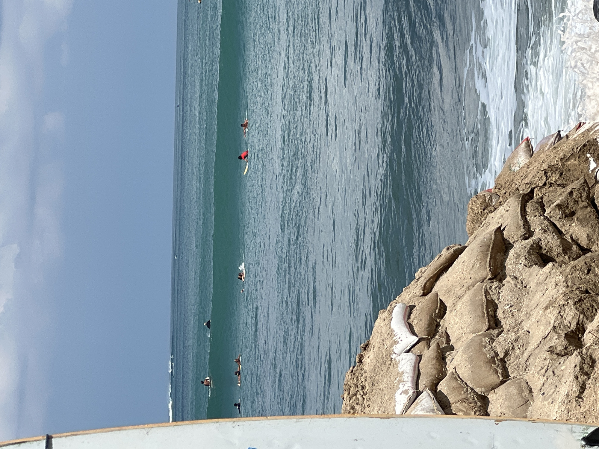 In Kuta, Indonesia, a stack of sandbags is piled on a rocky shore in the foreground, with a surfboard leaning against the left edge of the frame. The ocean stretches out to the horizon, with several surfers in the water, some on their boards and others paddling. The sky above is a mix of blue and clouds, and the water is a clear, turquoise color. The waves are breaking in the distance, creating a white foam line.