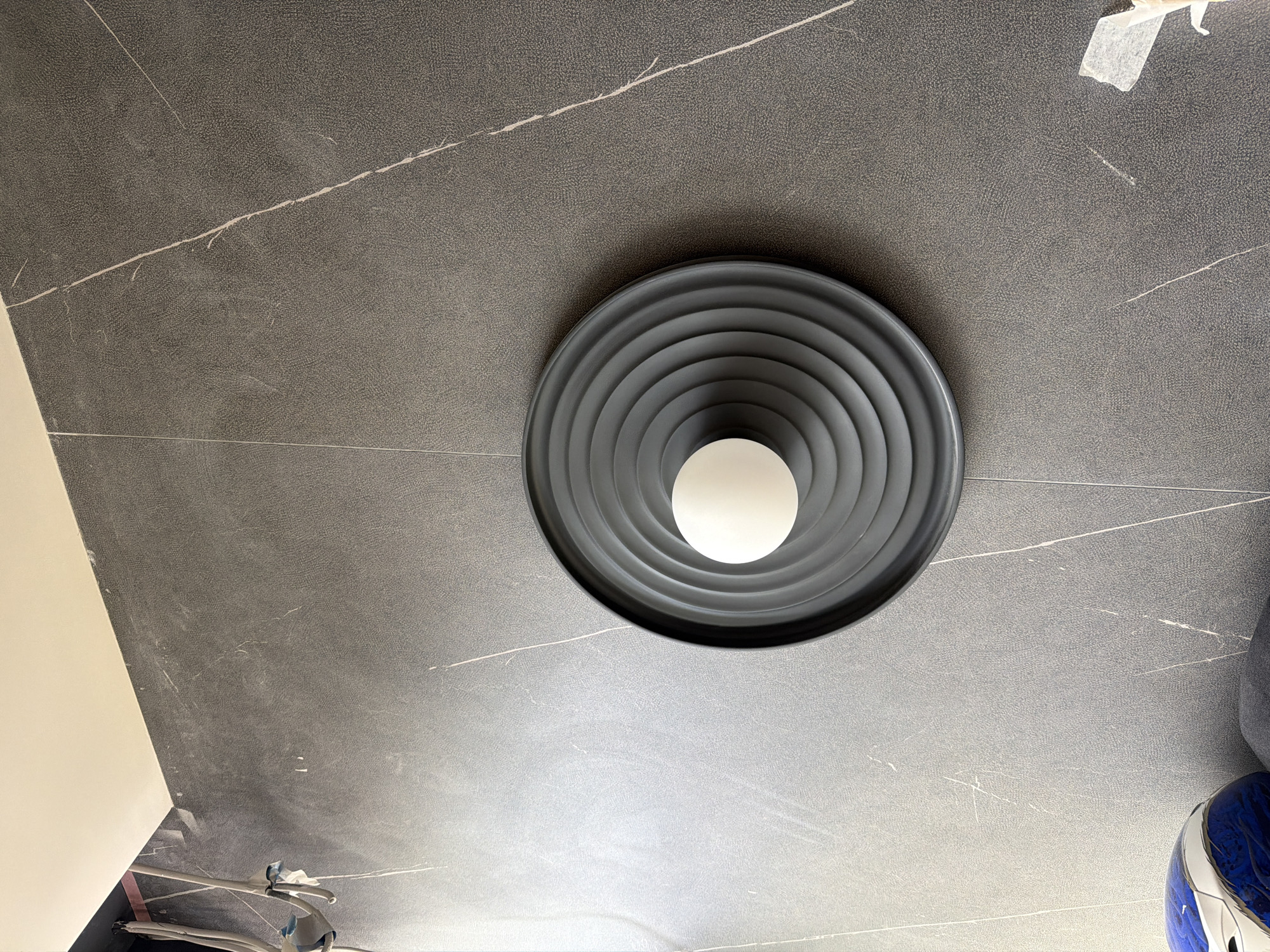 The image shows a low-angle shot of a ceiling light fixture set against a dark, textured ceiling with light-colored veining. The light fixture is round, gray, and has a series of concentric, circular ridges, with a spherical, white light source in the center. The ceiling appears to be made of tiles or large panels. The bottom corner of a white wall is visible in the lower-left of the frame.
The image does not contain sufficient information to determine a specific location within the city of Khimki, Russia, nor does it depict any people, actions, or interactions.