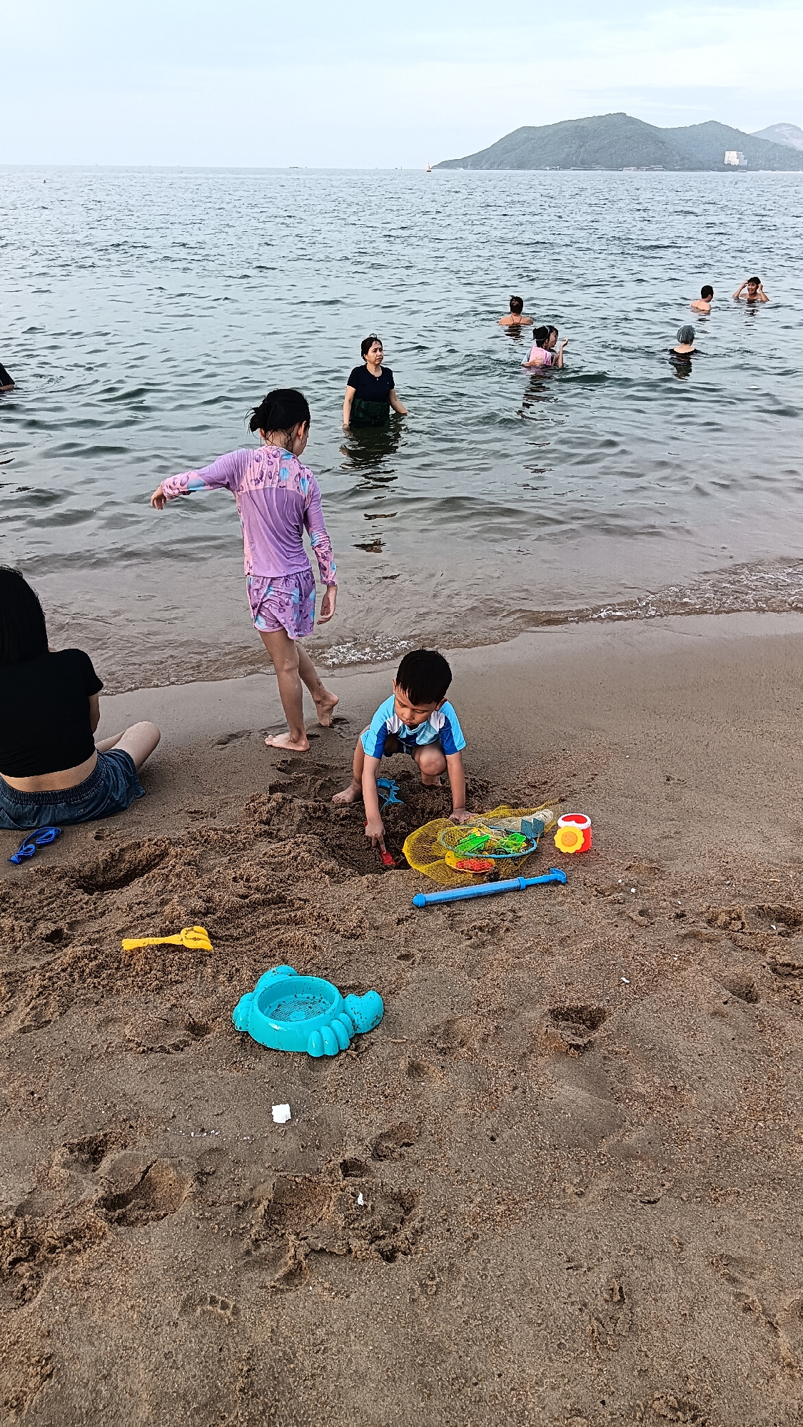 The image depicts a beach scene in Nha Trang, Vietnam, likely during the daytime with overcast skies. Several people are visible in the ocean, wading and swimming. On the sandy shore, a young boy is crouched down, playing with sand toys. He is wearing a blue and white striped shirt and dark shorts, and he is using a small red shovel. Next to him is a yellow mesh bag filled with sand toys, including a red and orange cylindrical toy and a blue toy. A blue plastic sand mold shaped like a turtle is also on the sand. A young girl in a purple swimsuit is standing near the water's edge, facing away from the camera. To the left, a woman in a black top and denim shorts is sitting on the sand, looking towards the water. The beach itself is composed of coarse sand with footprints and scattered debris. The ocean is calm with small waves lapping at the shore, and a distant coastline with hills is visible in the background.