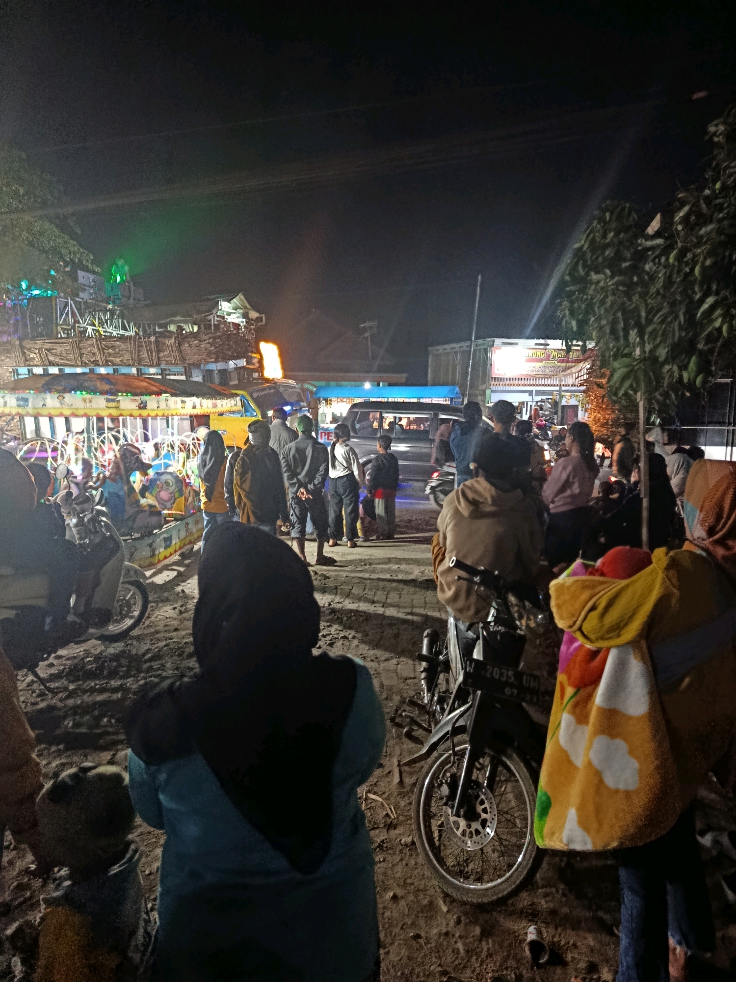In the city of Talangsuko, Indonesia, at night, a gathering of people is observed in an outdoor area, likely a market or public space. The scene is illuminated by artificial lights, including a large green spotlight and lights emanating from various stalls or attractions. In the foreground, a person wearing a teal headscarf and jacket faces away from the camera, with another person, partially visible, to their left. A child in a grey hoodie stands next to them. A parked motorcycle is visible in the mid-ground, with a colorful blanket draped over it.
Behind these individuals, a crowd of people are milling about. Some appear to be near a brightly lit, multi-colored vehicle or attraction, possibly a mobile carnival ride or food stall. In the background, more vehicles, including cars and motorcycles, are present, suggesting a busy thoroughfare or event. The ground is unpaved, appearing to be dirt or sand. The overall atmosphere is one of a community event or gathering taking place after dark.