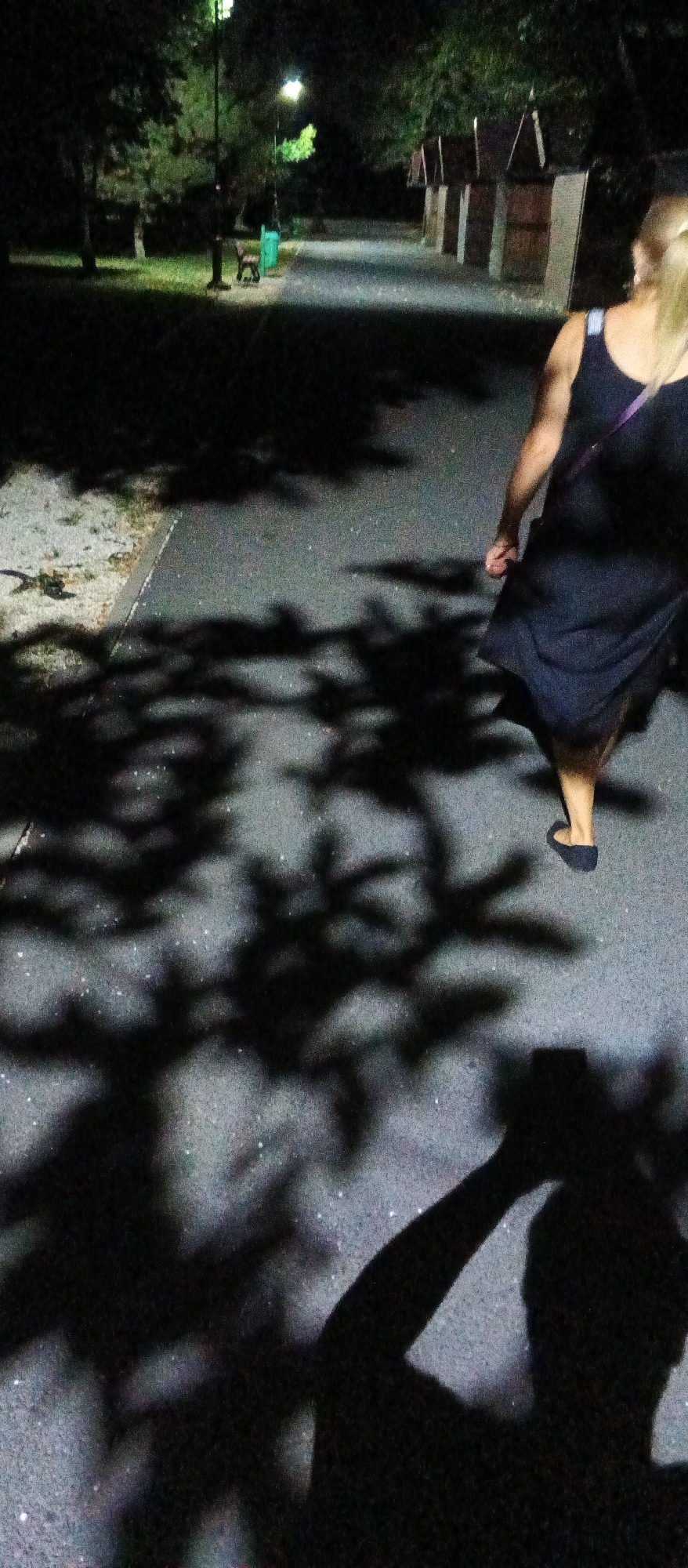 The image depicts an outdoor scene at night, possibly in a park or a recreational area in Mukachevo, Ukraine. In the foreground, the shadow of a person holding a smartphone is cast on the asphalt pathway. The person's arm is raised, suggesting they are taking a photograph or a video. Further along the pathway, a woman with blonde hair, wearing a dark dress and dark shoes, is walking away from the camera. Her shoulder bag strap is visible. The pathway is illuminated by a streetlamp, which also casts a shadow of tree leaves onto the ground. In the background, several small wooden structures, resembling booths or sheds, are lined up. Green grass and trees are visible to the left, and another streetlamp is present. A park bench and a green trash bin are also discernible near the pathway. The overall atmosphere is dimly lit, typical of a nighttime setting.