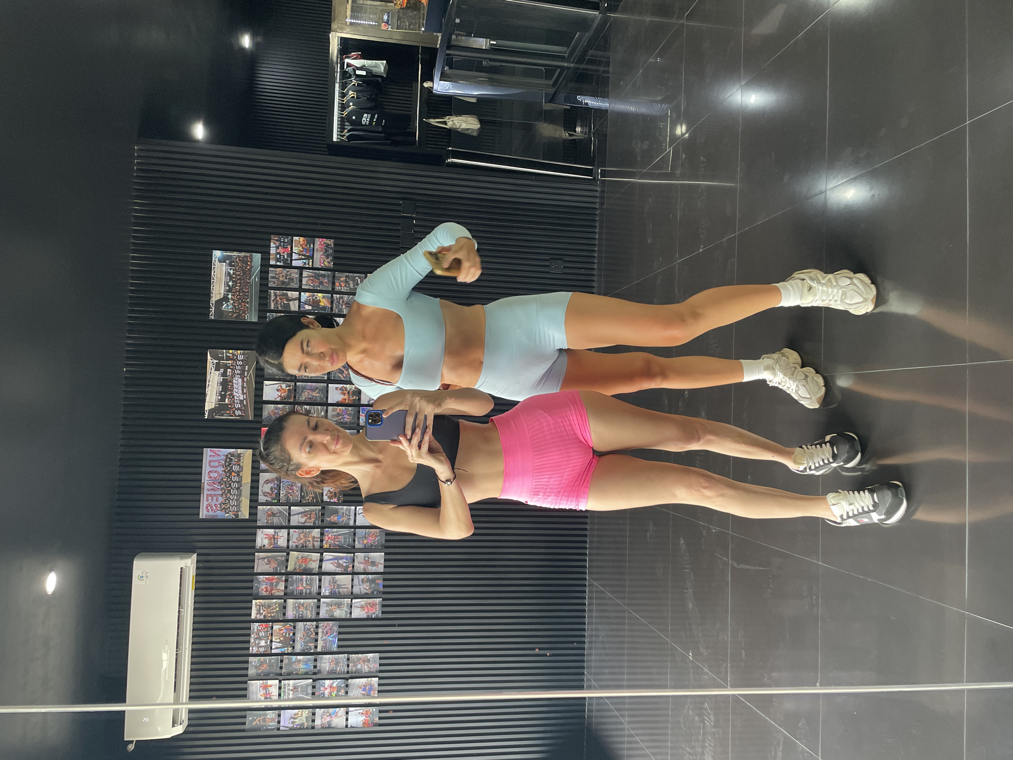 Two women are posing for a mirror selfie in a gym. The woman on the left is wearing a black sports bra and bright pink shorts. She is holding a phone in her hands and looking at her reflection. The woman on the right is wearing a light blue long-sleeved crop top and matching light blue shorts. She is holding a phone in her right hand and looking at her reflection with a playful expression. They are both wearing athletic shoes. The gym has a modern aesthetic with black paneled walls and a glossy black tiled floor. There are framed photos displayed on the wall behind them. An air conditioning unit is mounted on the wall above the photos. The overall environment suggests a fitness center in Mengwi, Indonesia.