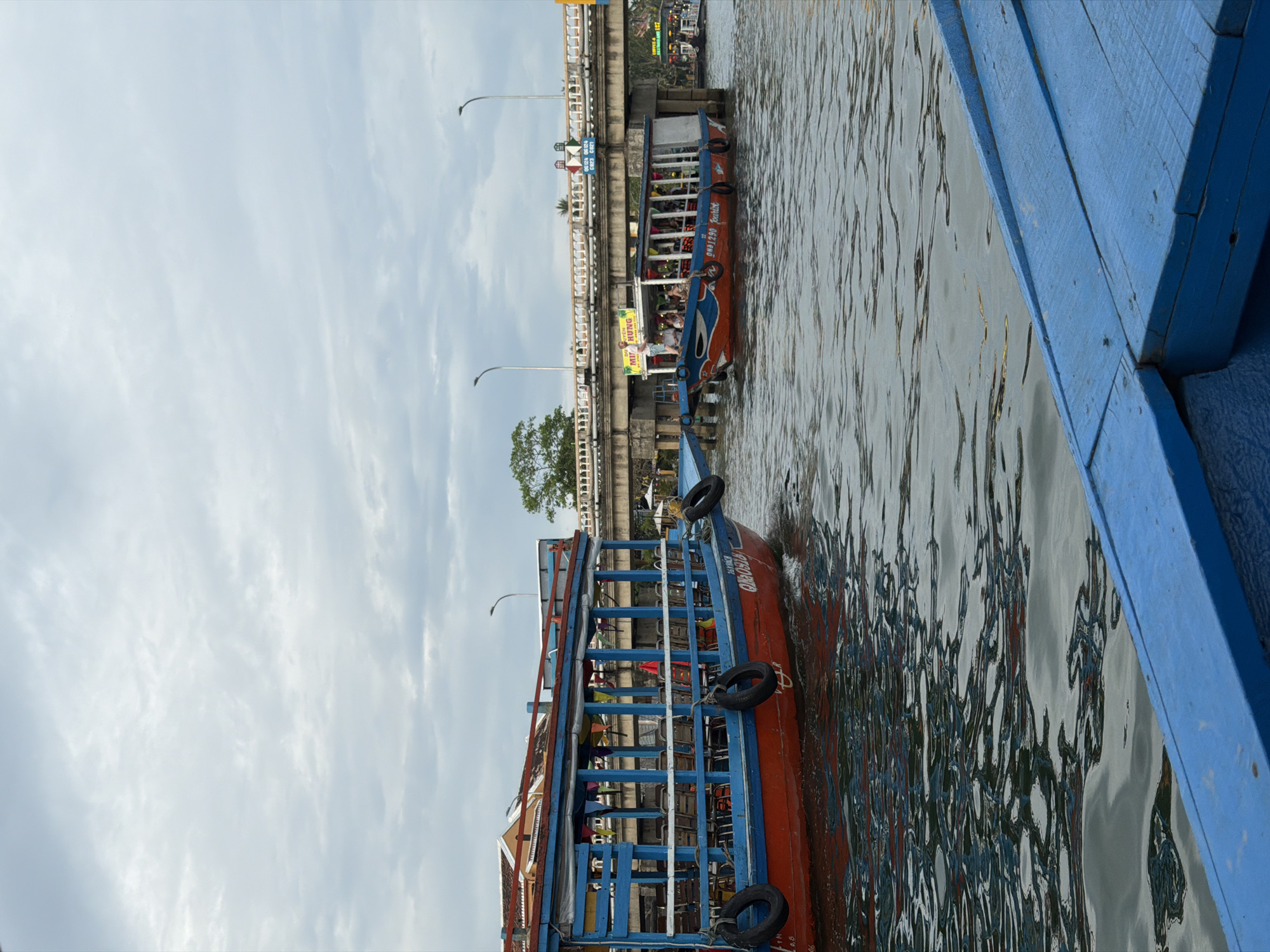 This image captures a vibrant scene along a river or canal in Hội An Ward, Vietnam, featuring boats and a bridge under an overcast sky. The photograph is oriented vertically.
In the immediate foreground, partially visible on the right, is a blue wooden structure, likely the side of another boat from which the photo was taken. The water surface next to it shows gentle ripples and reflections.
A prominent feature in the mid-ground is a passenger boat with a red hull and blue railings, carrying numerous people. Black rubber fenders are attached to its sides. Visible text on the boat includes "QN01290" and the word "Dung." This boat appears to be moving or docked close to a concrete bridge.
The bridge, which spans across the waterway, is equipped with white railings, streetlights, and various signs. On the underside of the bridge, a yellow sign advertises "MÌ HÙNG" (Mi Hung) along with a phone number, "0935 329...". Another blue sign, partially visible, reads "CẦU AN HỘI," indicating it is the An Hoi Bridge. People can be seen walking and standing on the bridge.
Beyond the bridge, the river continues, with glimpses of distant buildings and other boats. A few green trees are also visible near the bridge's support. The sky above is cloudy and uniformly light grey, suggesting an overcast day. The scene conveys a busy, functional waterscape, characteristic of a town with riverine activity, consistent with the provided location context of Hội An.