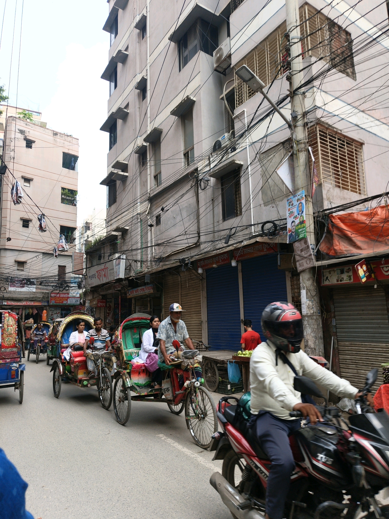 The image depicts a street scene in Dhaka, Bangladesh, characterized by dense urban architecture and a busy thoroughfare. In the foreground, a person wearing a white shirt, dark pants, and a red and black helmet is riding a motorcycle. Behind the motorcycle, several cycle rickshaws are present, some carrying passengers. One rickshaw features a woman in a white outfit and a man in a striped shirt. Another rickshaw, partly obscured, shows a man in a blue shirt. Further in the background, more rickshaws and buildings line the street. The buildings are multi-story structures with visible windows and a complex network of electrical wires suspended overhead, a common feature in Dhaka's urban landscape. The street is paved with asphalt, and the overall atmosphere suggests a lively, densely populated urban environment.