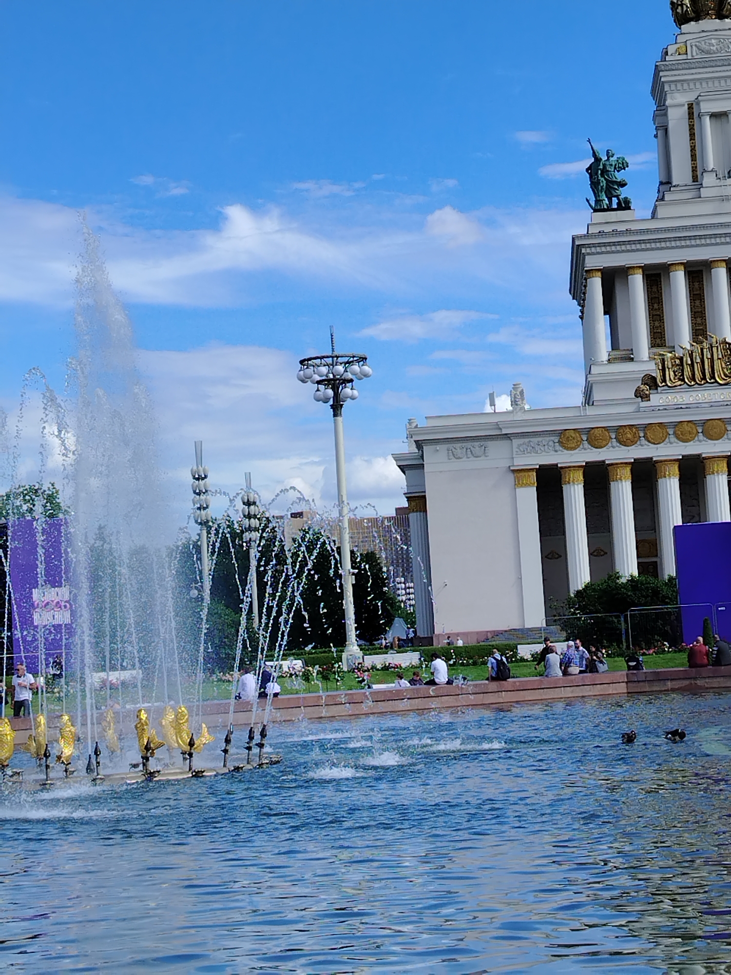 This image captures a sunny day in Moscow, Russia, likely in the late morning or early afternoon, given the bright lighting and clear blue sky with scattered clouds. The scene features a large, ornate fountain in the foreground, with water jets spraying high into the air. The fountain has several golden statues at its base. In the background, a grand, neoclassical building with white columns and golden accents stands tall. A sculpture of two figures adorns the top of the building.
Several people are scattered around the area, some sitting on the grass near the fountain, while others are walking or standing. The overall atmosphere appears relaxed and pleasant, suggesting a leisurely day out. To the left, a large purple banner with some text is visible, though the text is not clearly legible. The presence of trees and greenery indicates a park or public square setting. The architecture of the building strongly suggests the location is VDNH (Exhibition of Achievements of National Economy), a prominent exhibition center and park in Moscow known for its fountains and grand pavilions.