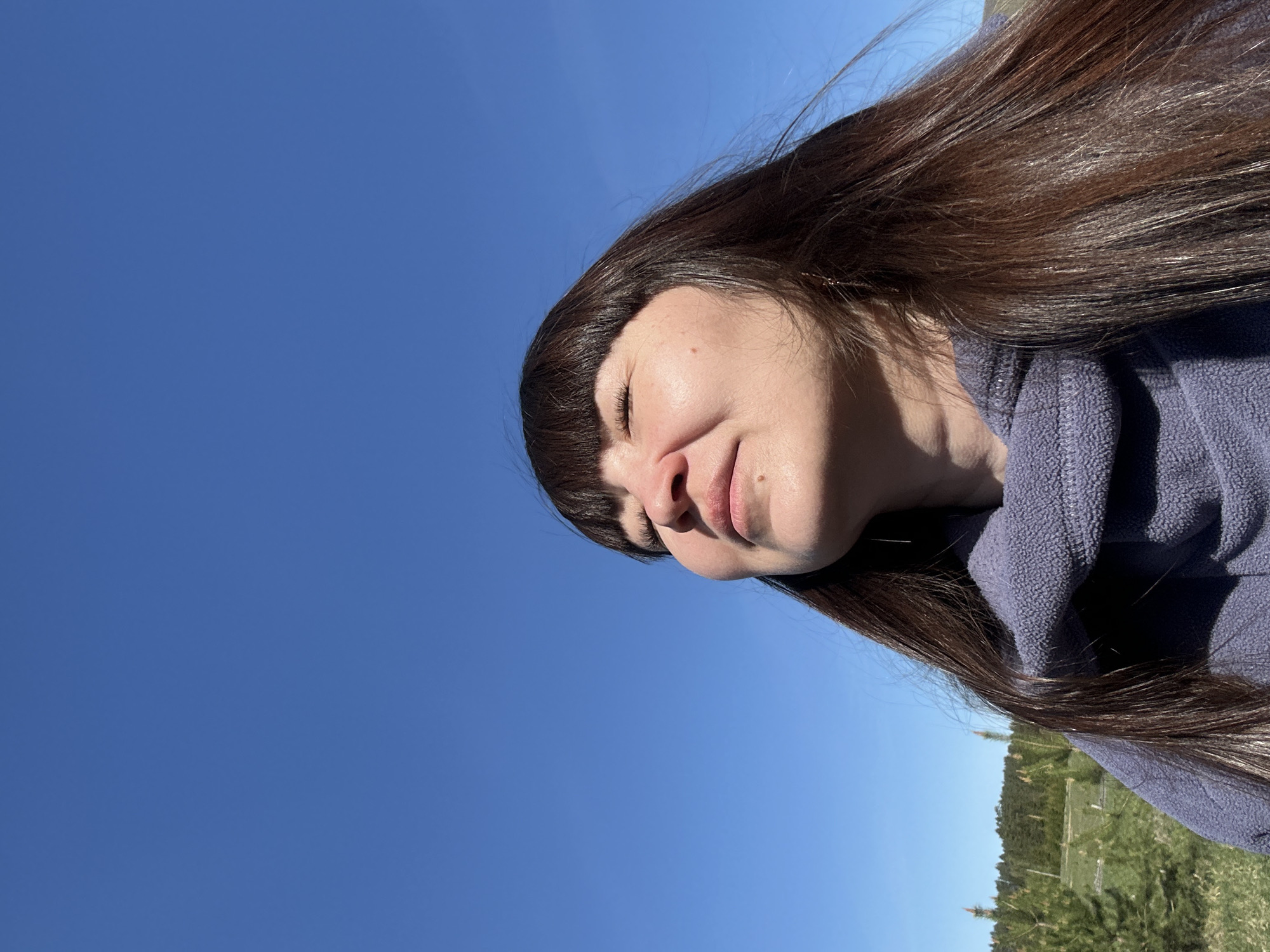 A woman with long, dark brown hair and bangs is captured in a close-up shot against a backdrop of clear blue sky. She has her eyes closed and her head is tilted back slightly. She is wearing a blue fleece jacket. The environment includes trees and vegetation in the background, suggesting an outdoor setting possibly in Среднее Кечево, Russia.