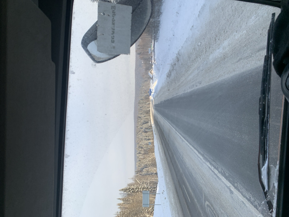 The scene depicts a snow-covered highway in a rural area outside the city of Div'ya, Russia, viewed from inside a vehicle. The asphalt road, partially cleared of snow and ice, extends straight into the distance, flanked by substantial snow banks. Dense evergreen forests, heavily laden with snow, line both sides of the road. The sky is bright and overcast. On the left side of the road, two blue-and-white road signs are visible within the snow-covered verge. On the right side, a small wooden building with a snow-covered roof and other minor structures are visible beyond the snow bank. Within the vehicle's interior, the dashboard and wiper blades are visible at the bottom of the frame. A rearview mirror is partially visible in the upper right quadrant, from which a rectangular tag displaying reversed text, possibly "ЕБОВКУ93," is suspended. A gloved hand is visible in the bottom right corner of the frame.